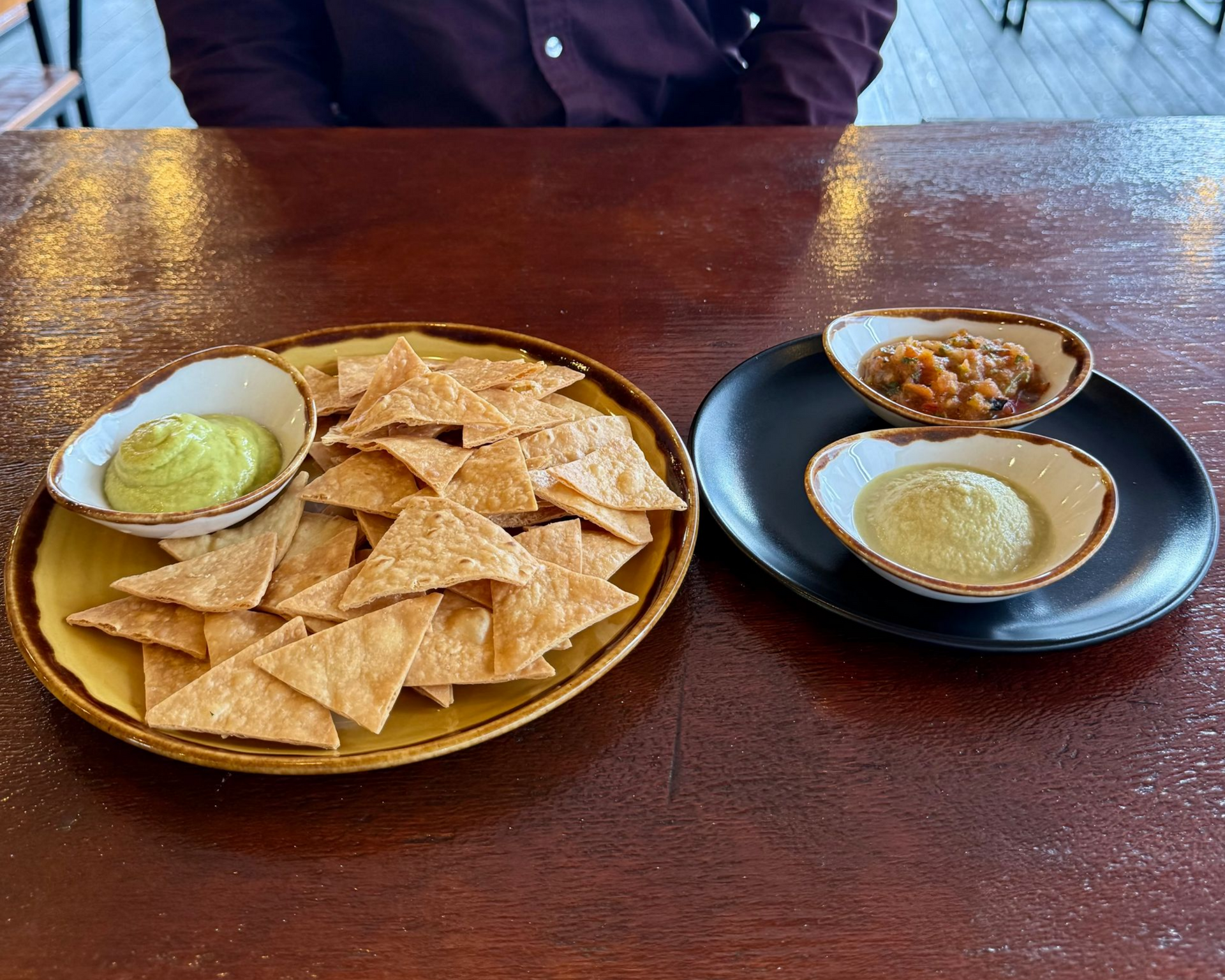 Tortilla chips with guacamole and bean dips on a wooden table. A person is in the background.