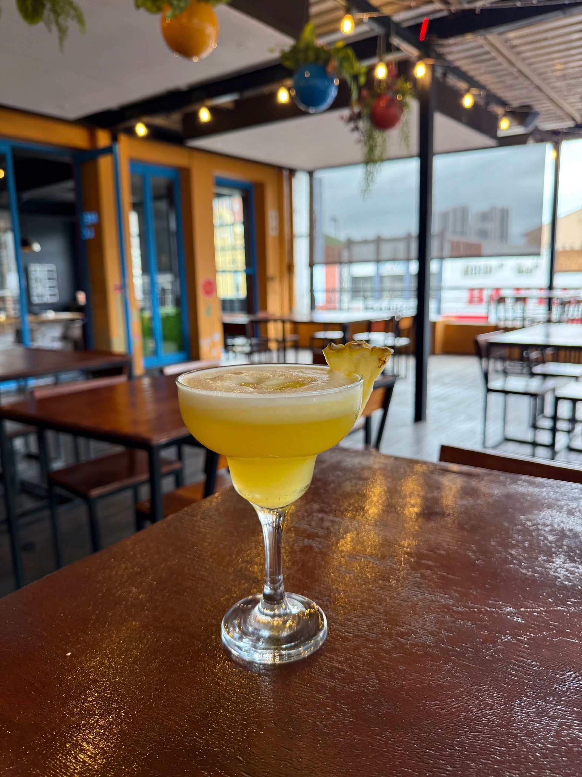 A yellow cocktail in a stemmed glass on a wooden table, with an outdoor cafe background.