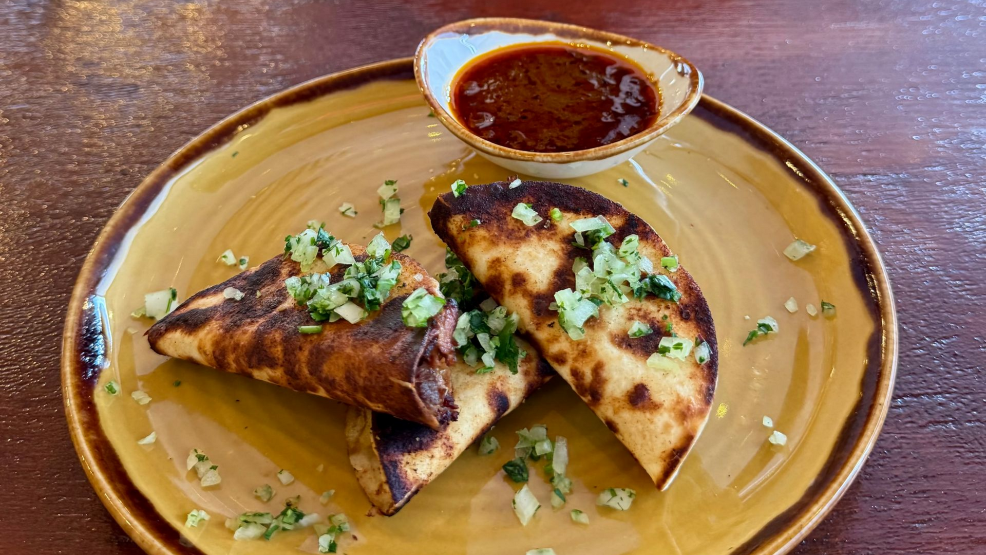 Three tacos on a plate with sauce. Tacos are golden brown with green topping, served with red sauce.