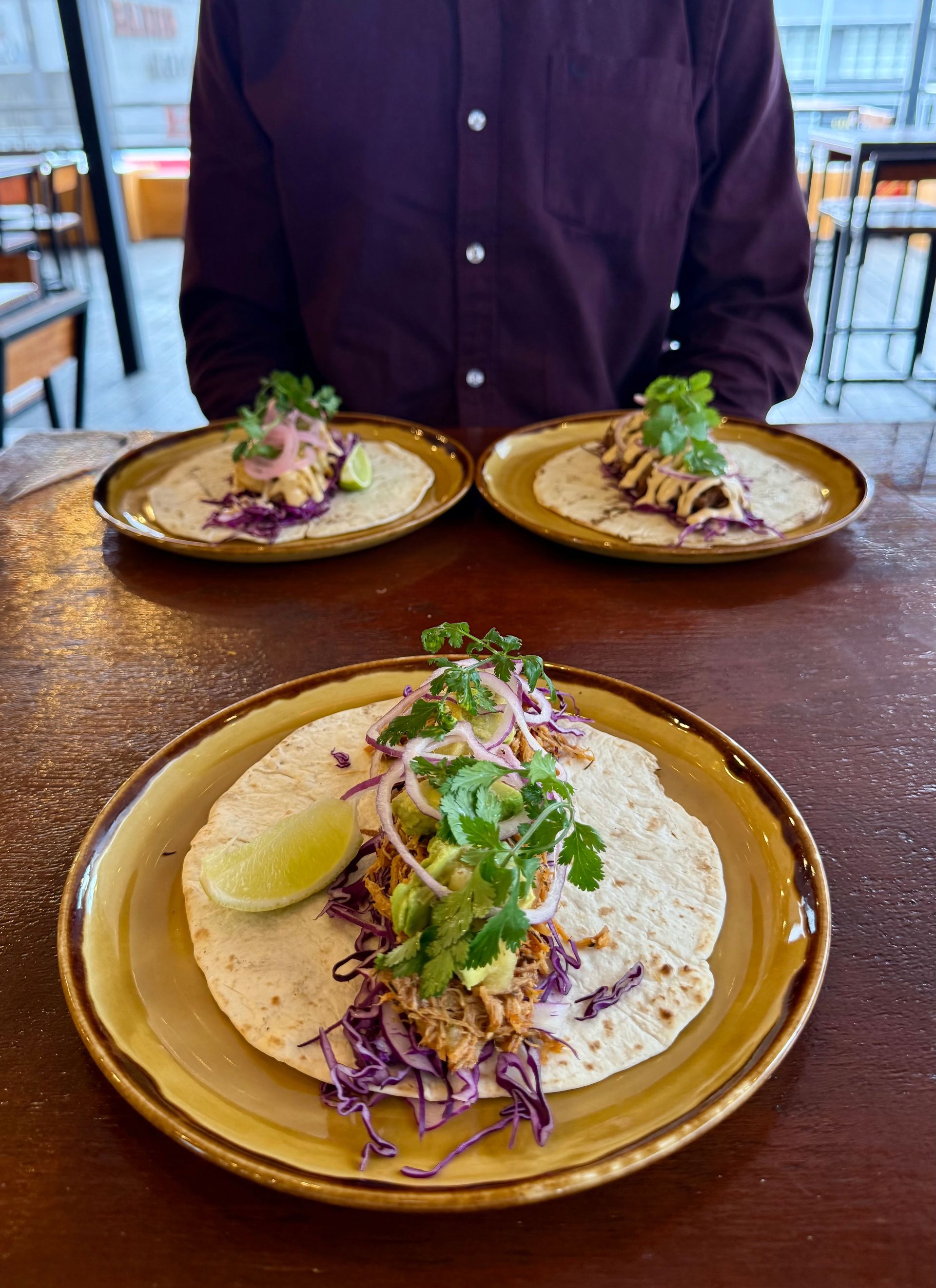Three tacos on plates with toppings and a lime wedge, served on a table.