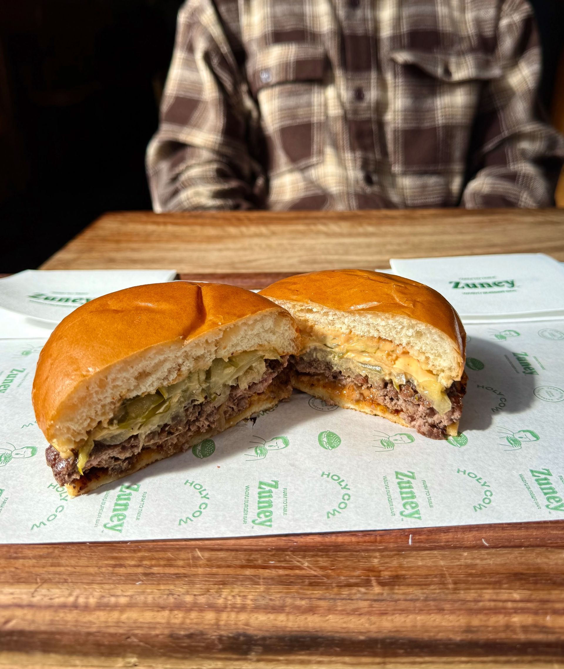 A sliced hamburger on a wooden board, with a person in a plaid shirt in the background.