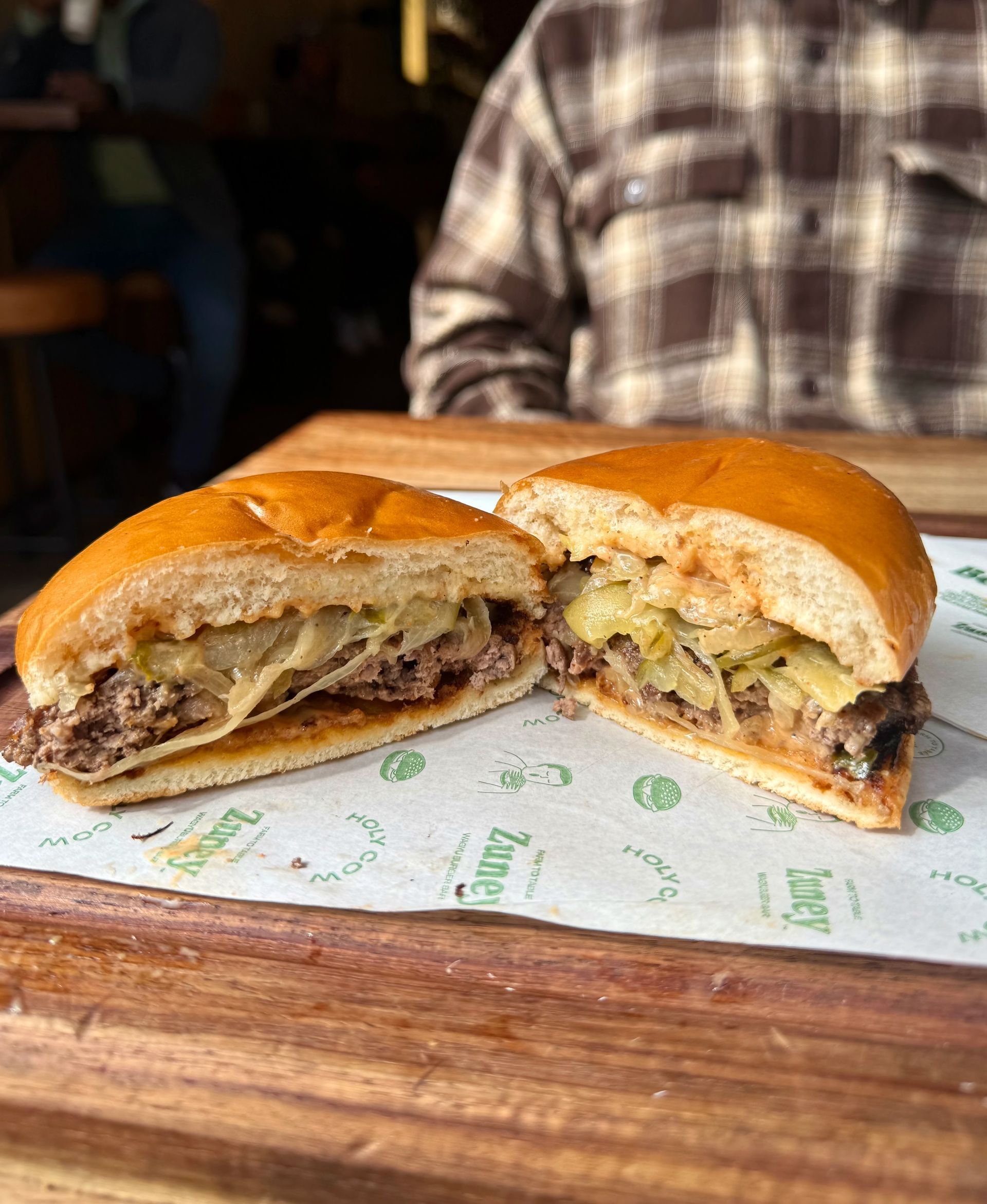 A halved cheesesteak sandwich on a wooden board. The sandwich sits on printed paper; person in plaid shirt in the background.