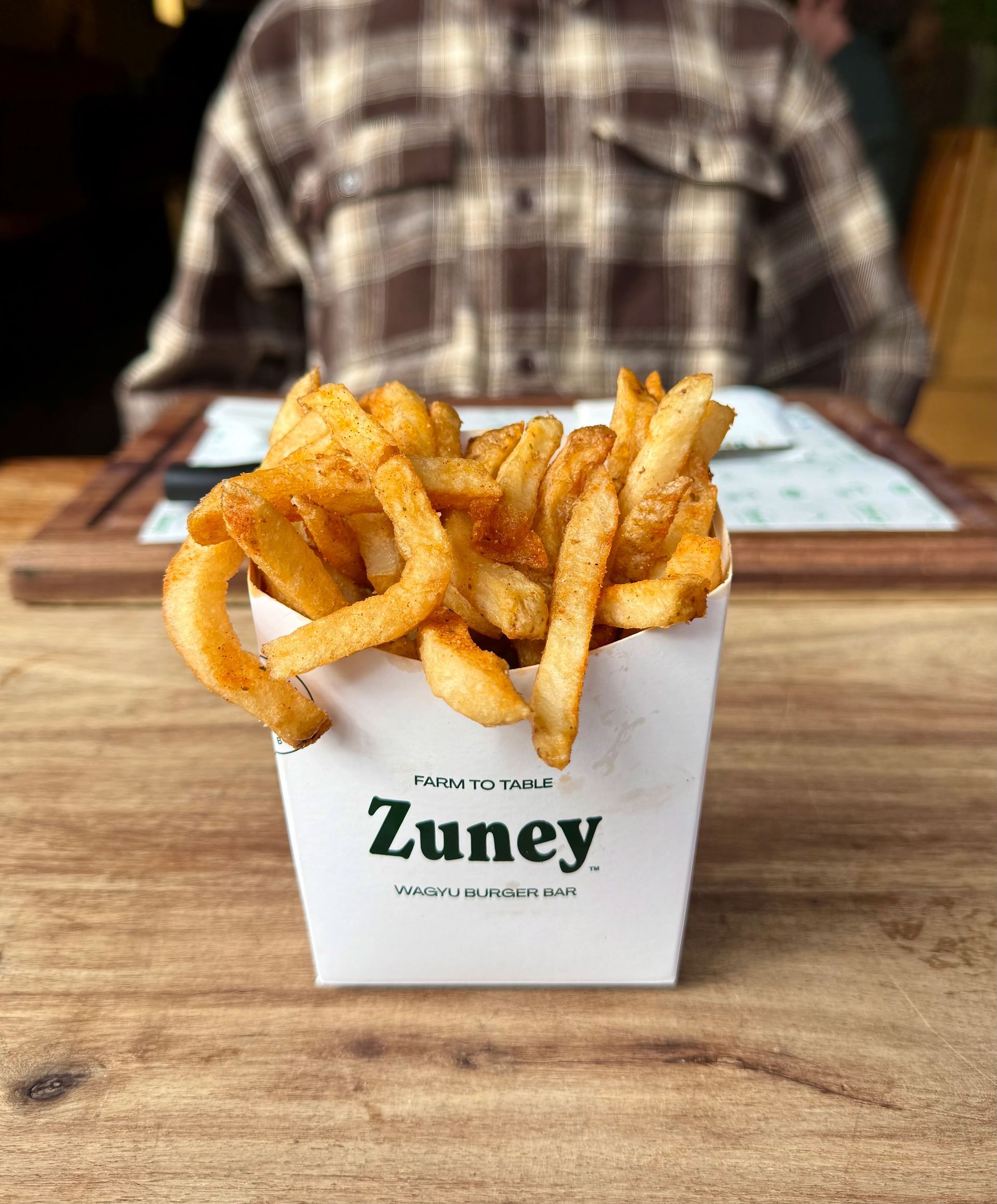 Fries in a white paper container on a wooden table, person in flannel shirt in background.