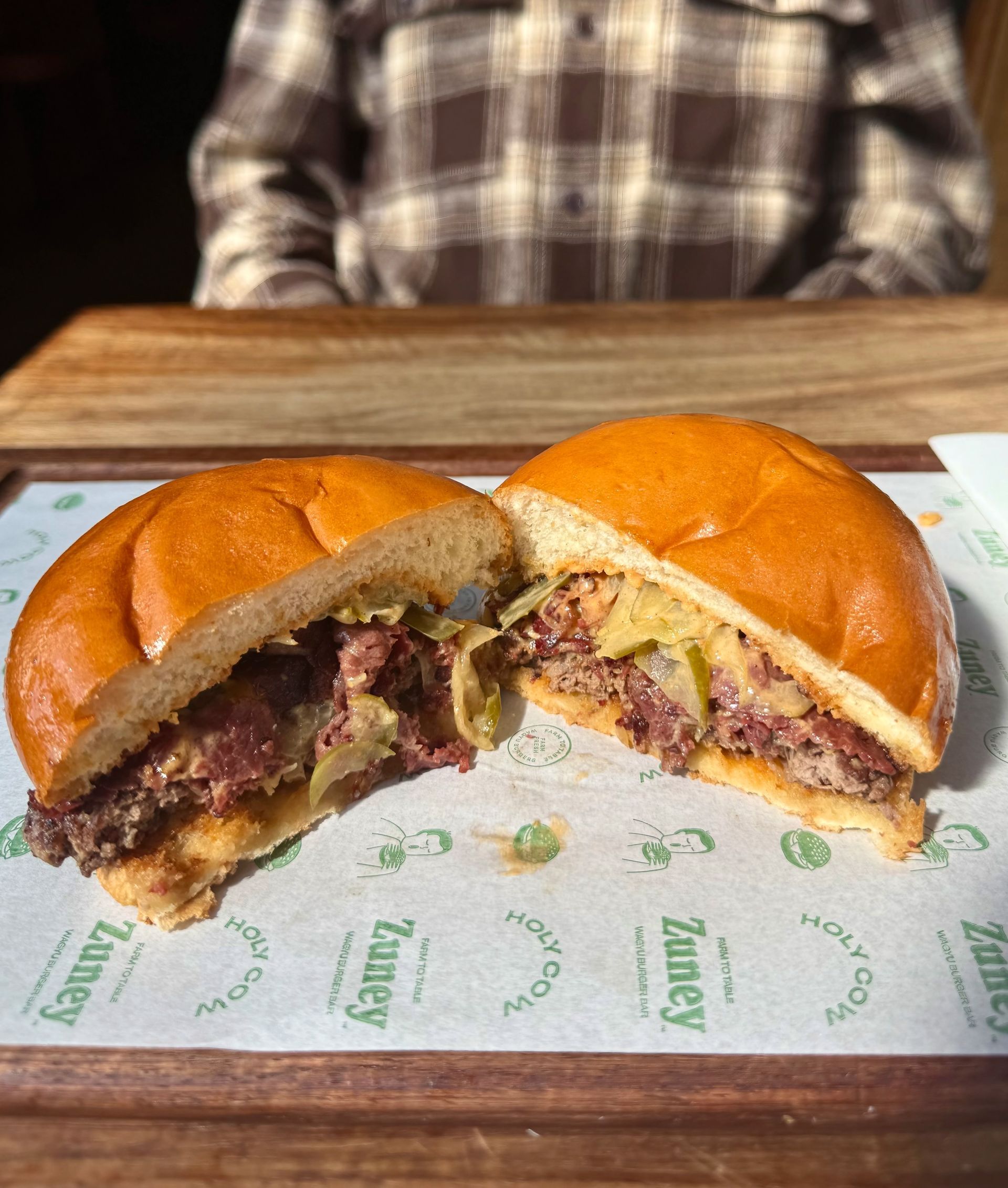 A halved burger on a branded paper, brown patty and bun, yellow cheese and pickles.
