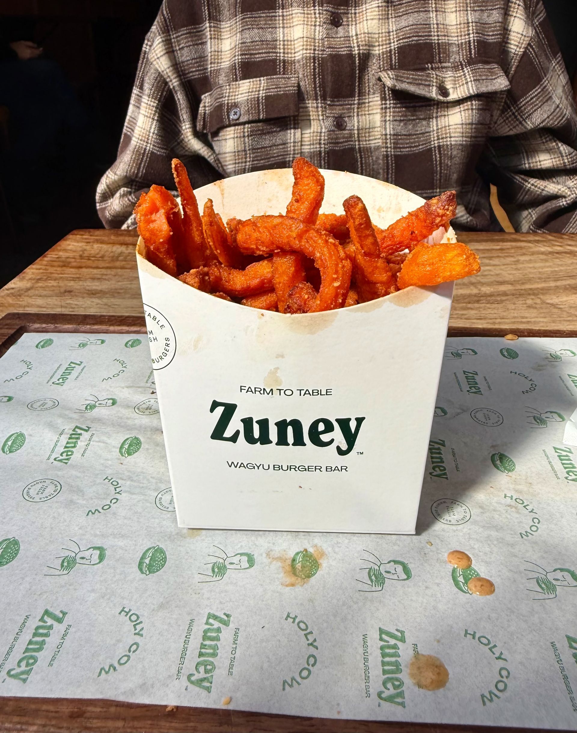 Sweet potato fries in a Zuney branded container on a table with someone wearing a plaid shirt.