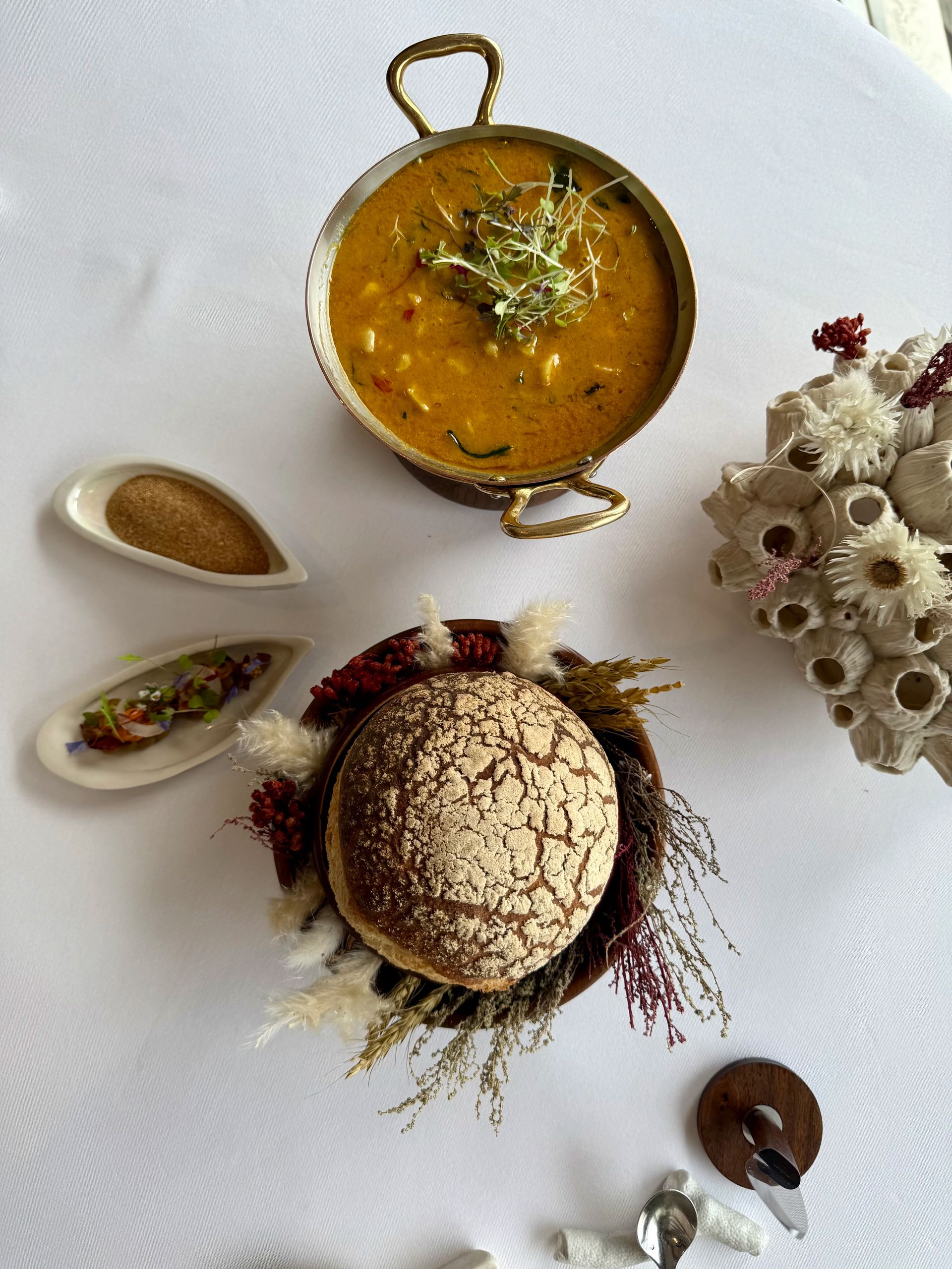 A golden-brown soup in a handled bowl with bread and spices on a white tablecloth.