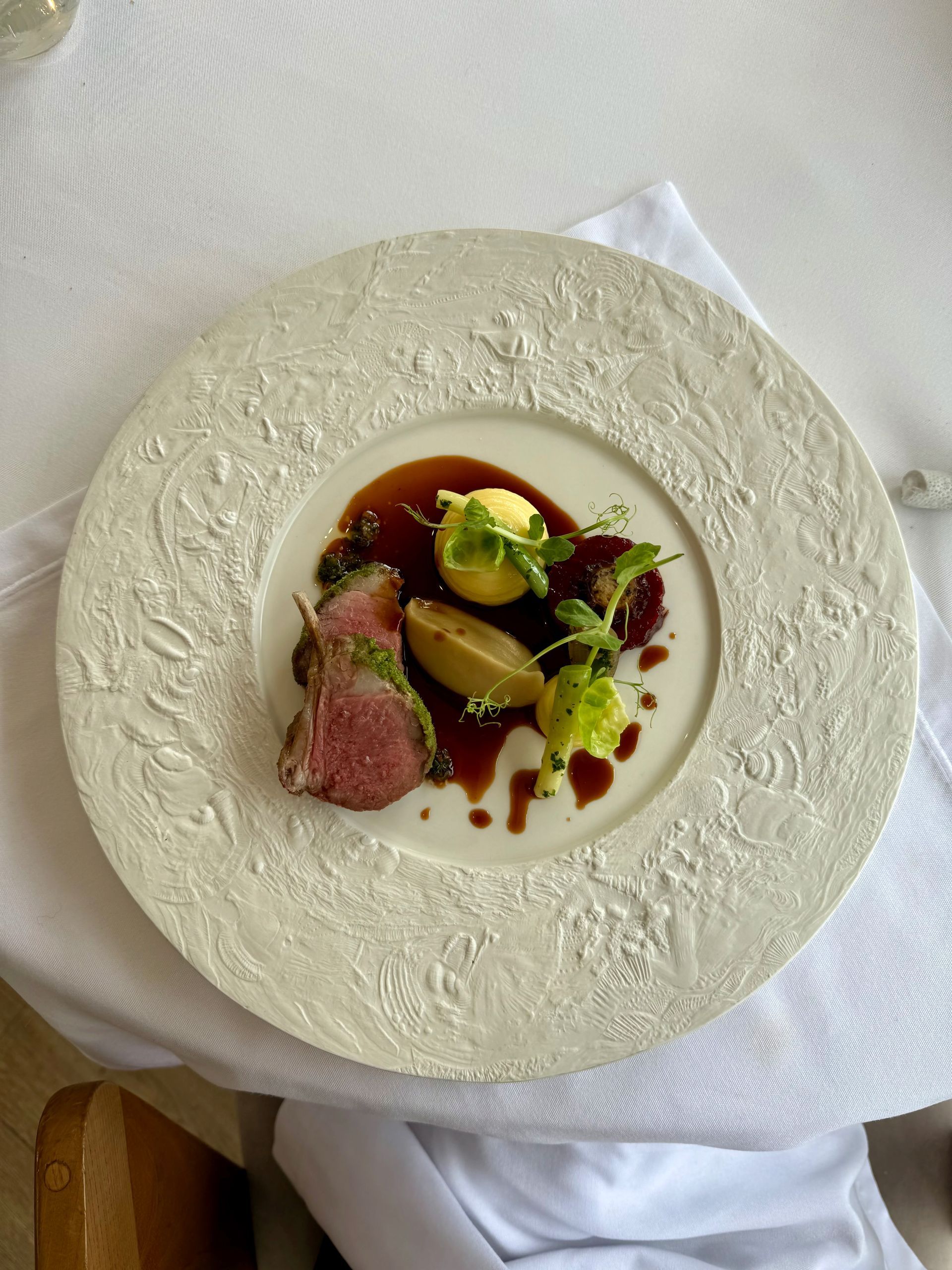 Roasted lamb with sauce, potatoes, and garnish on a patterned white plate.
