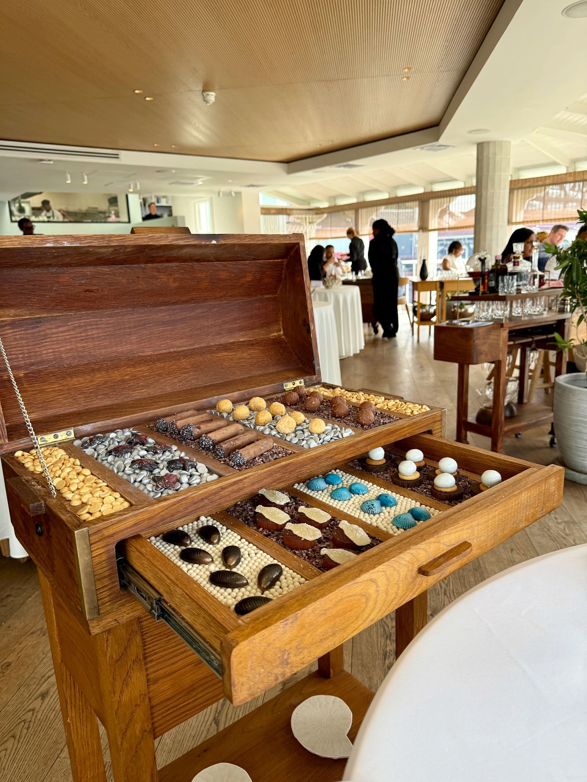 Wooden chest with various chocolates in a restaurant setting, with people visible.