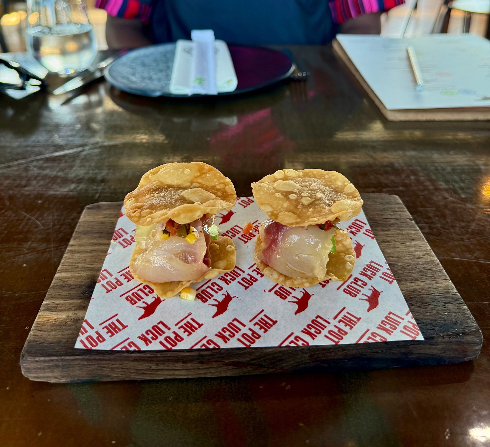 Two sushi appetizers on a wooden board; topped with crispy wonton, served on branded paper.