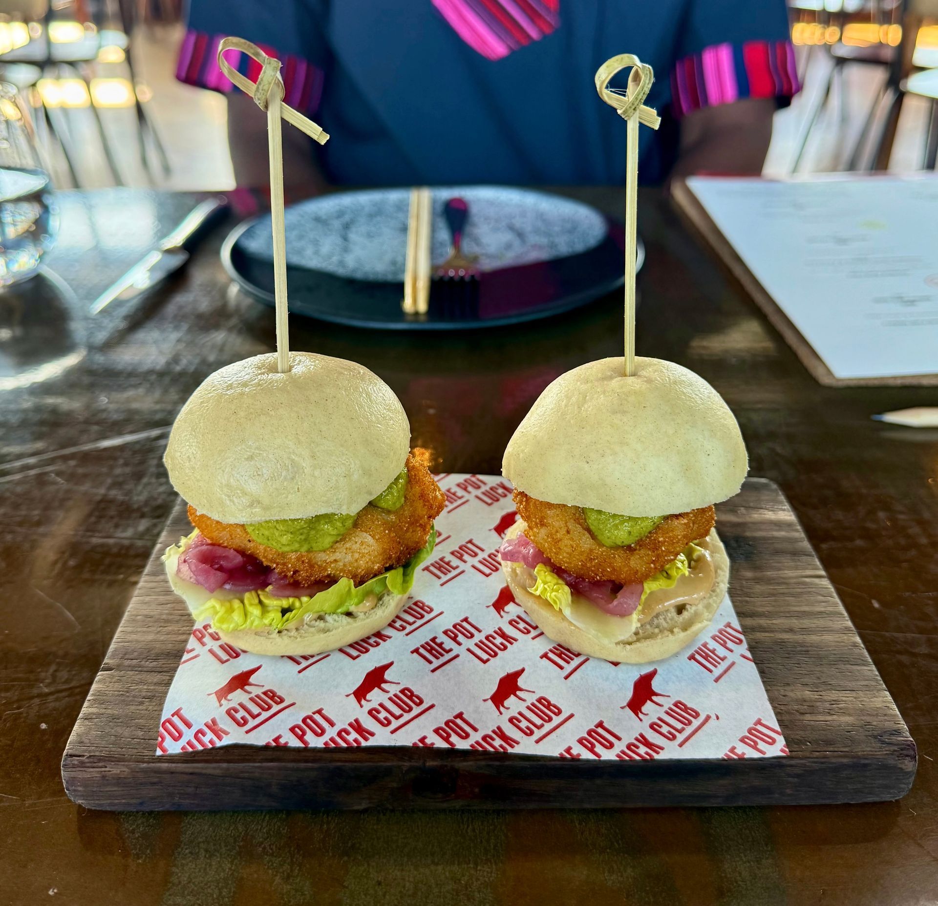 Two sliders on a wooden board with pickles, onions and sauces. Pot Lock Club logo on paper.