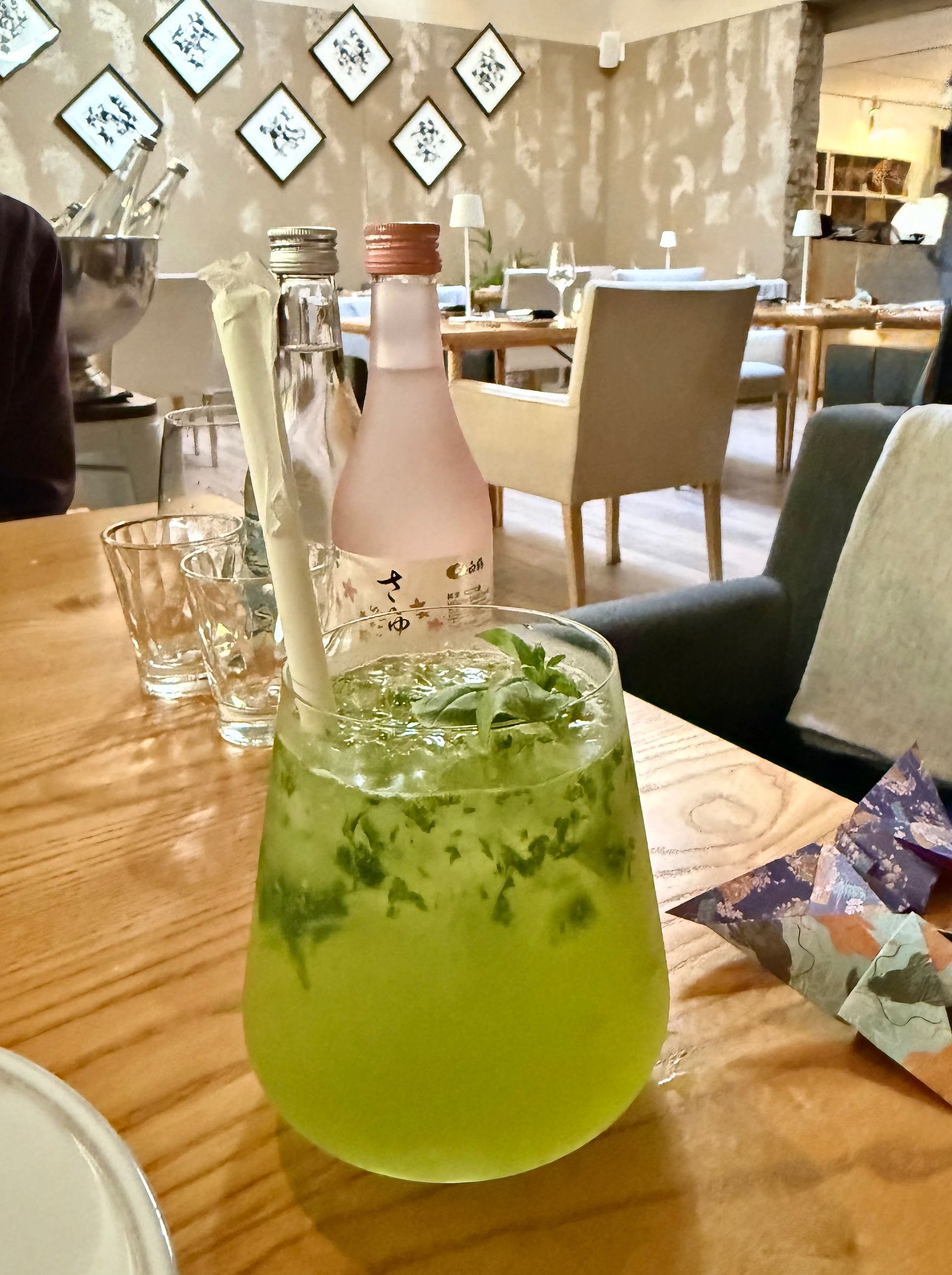 A green cocktail with a paper straw sits on a wooden table in a restaurant.