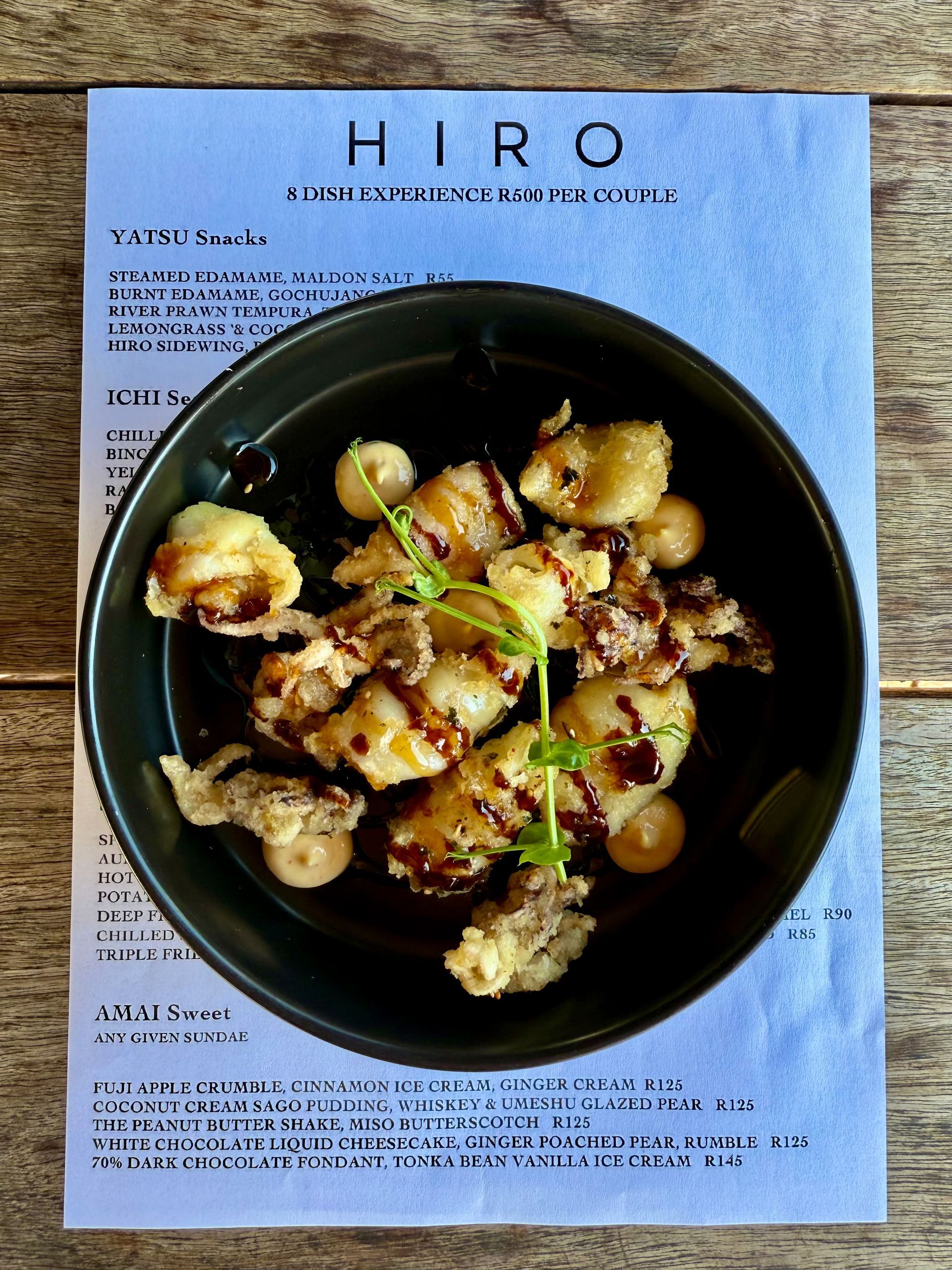 Fried dish in a dark bowl, drizzled sauce, atop a menu, and a wooden table.