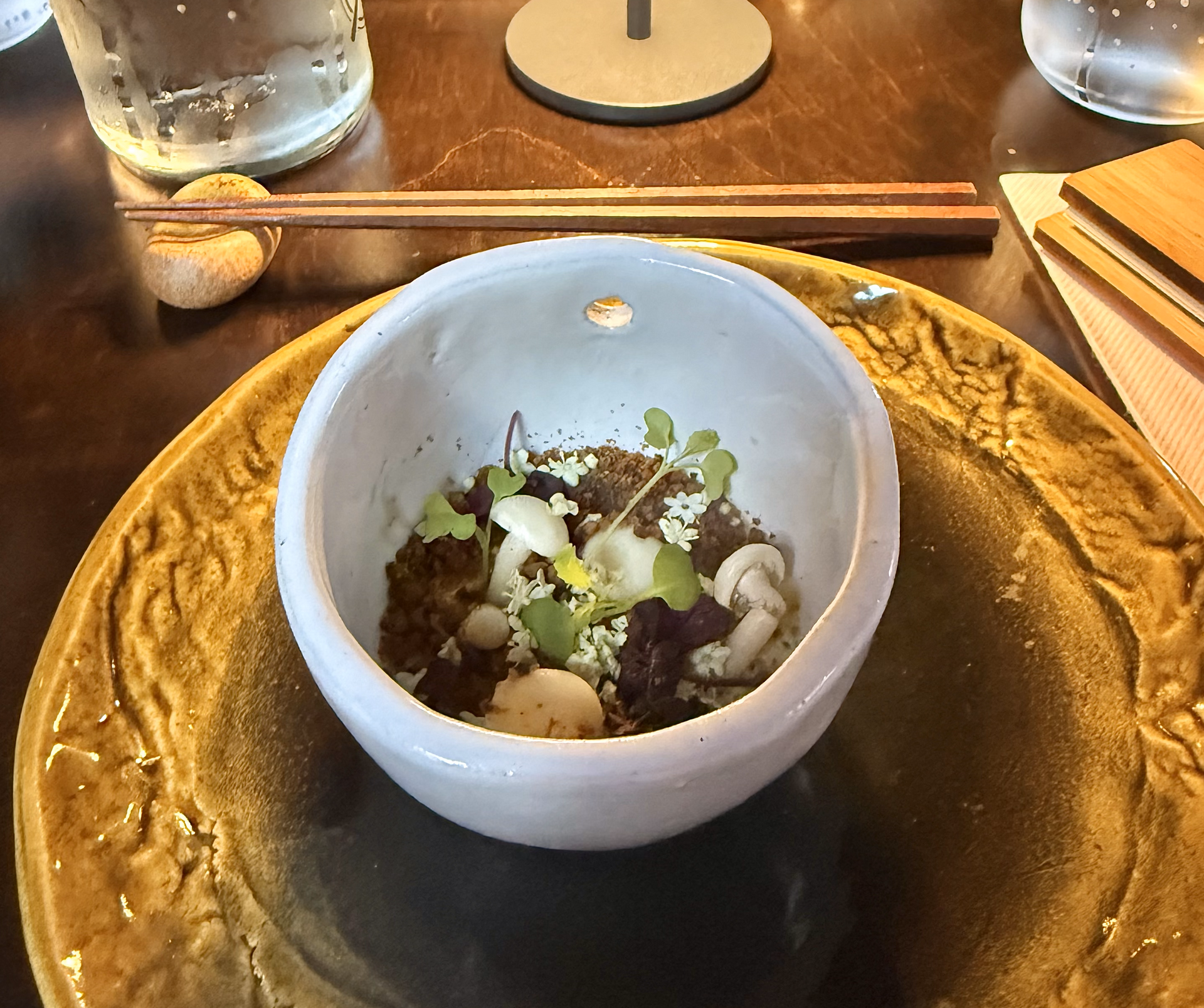 Small bowl of food on a gold-colored plate with chopsticks, a water glass, and wood table in the background.