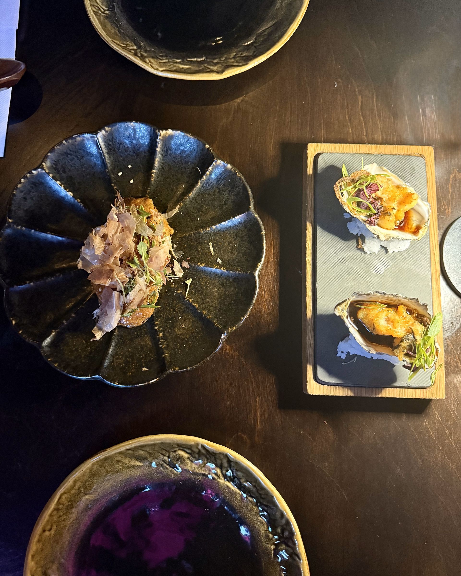 Dark wooden table with three decorative plates holding food; two oysters, and a meat dish.