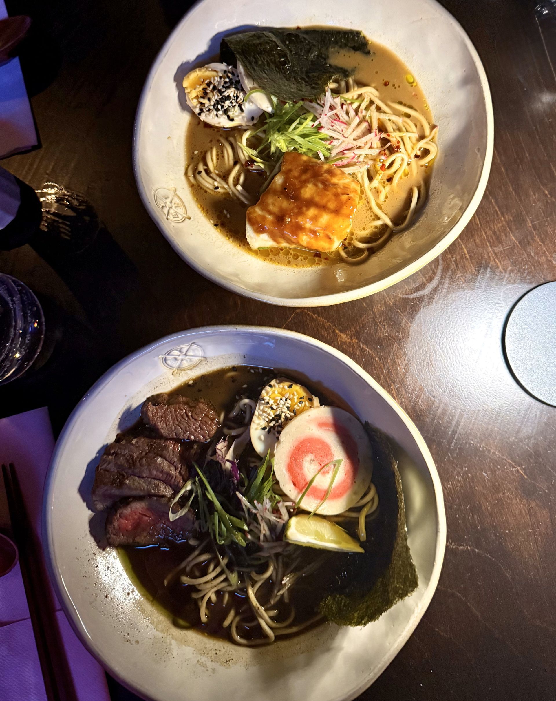 Two bowls of ramen with various toppings on a dark wooden table.