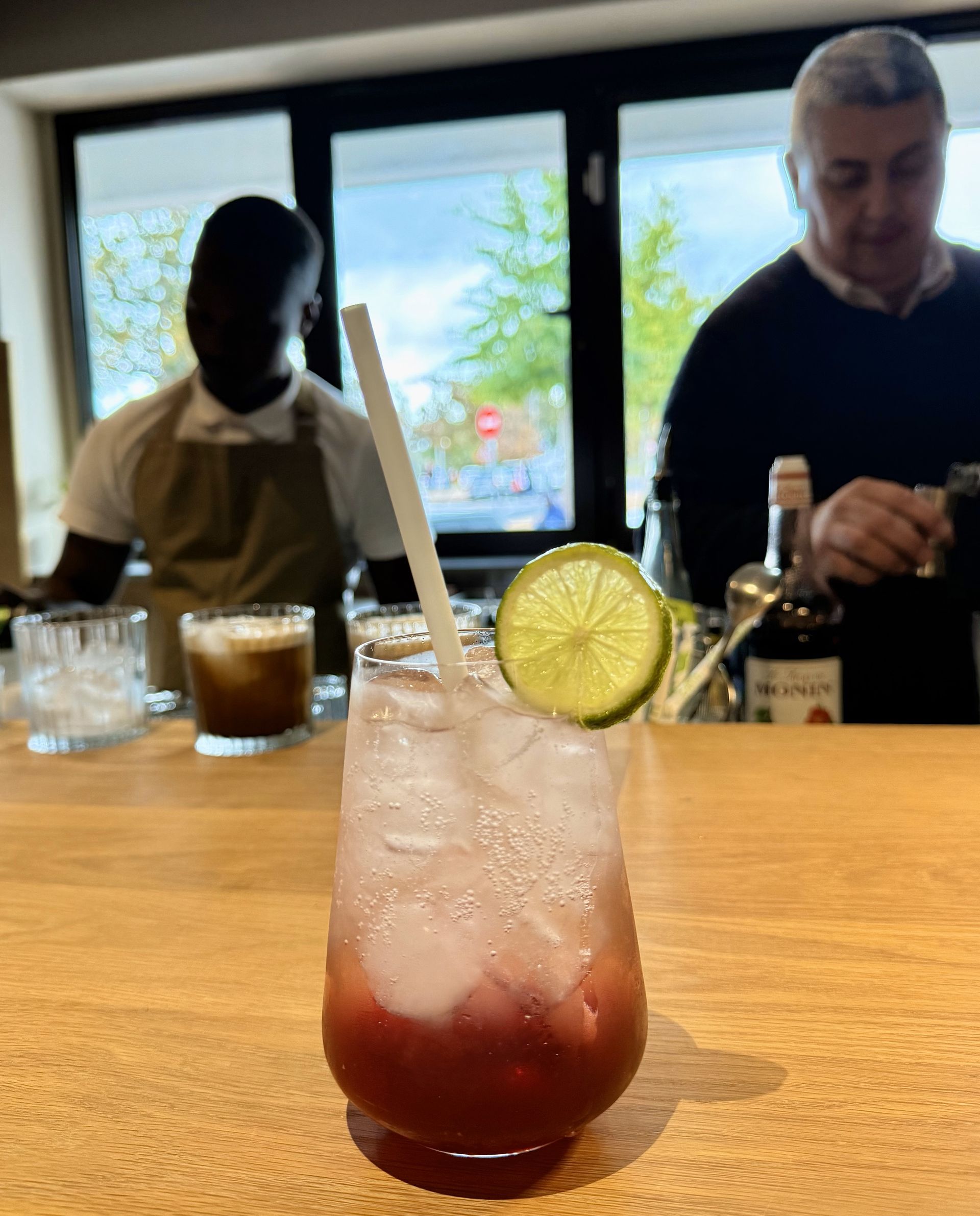 Cocktail with lime and straw on a bar, with two bartenders in the background.