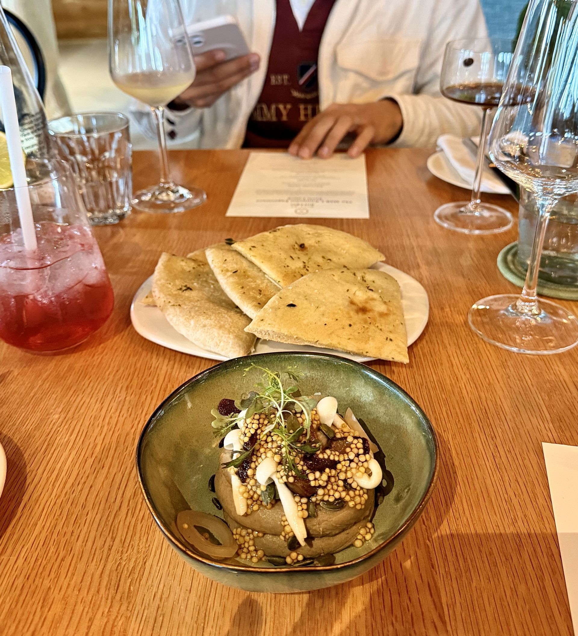 A table set with food, wine, and a drink. A person in the background.