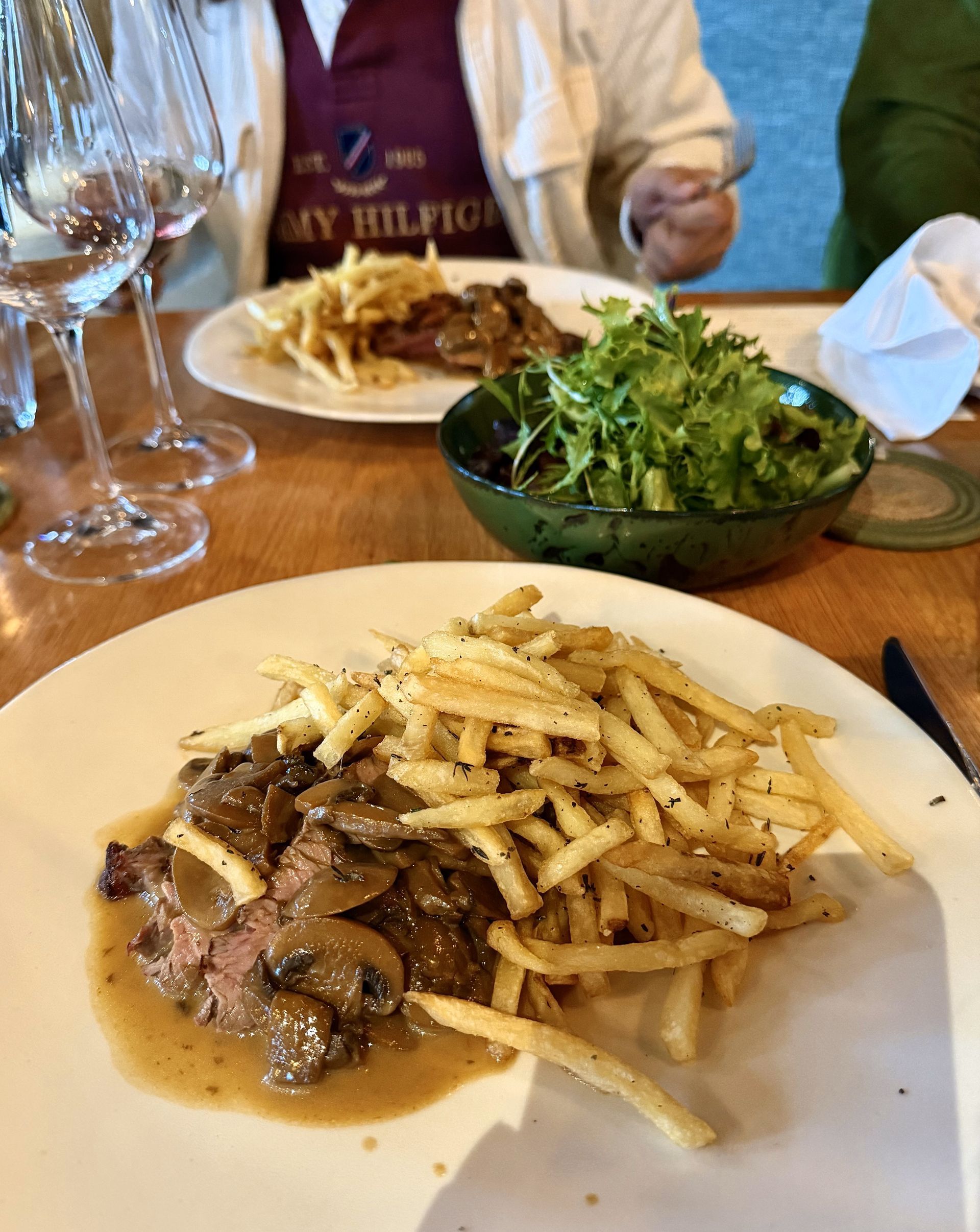 Steak with mushroom gravy and fries on a white plate, with a salad and another plate of food in the background.