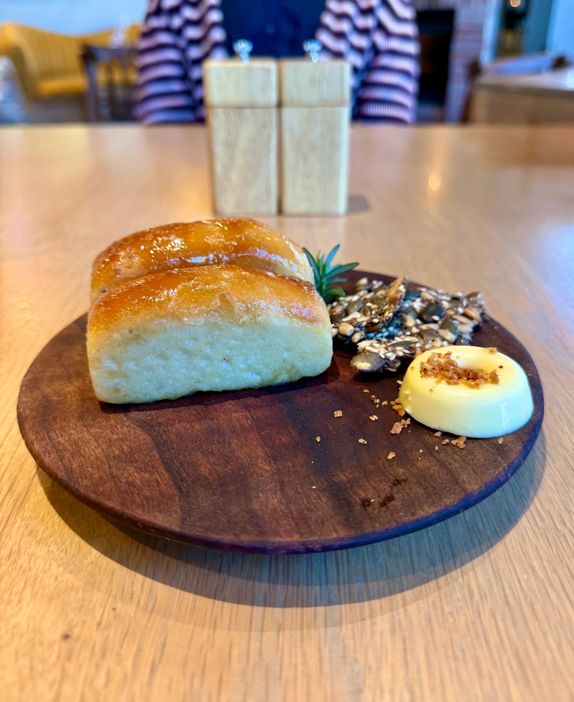 Roll, seeds, and butter on a wooden plate. Salt and pepper shakers and person in background.