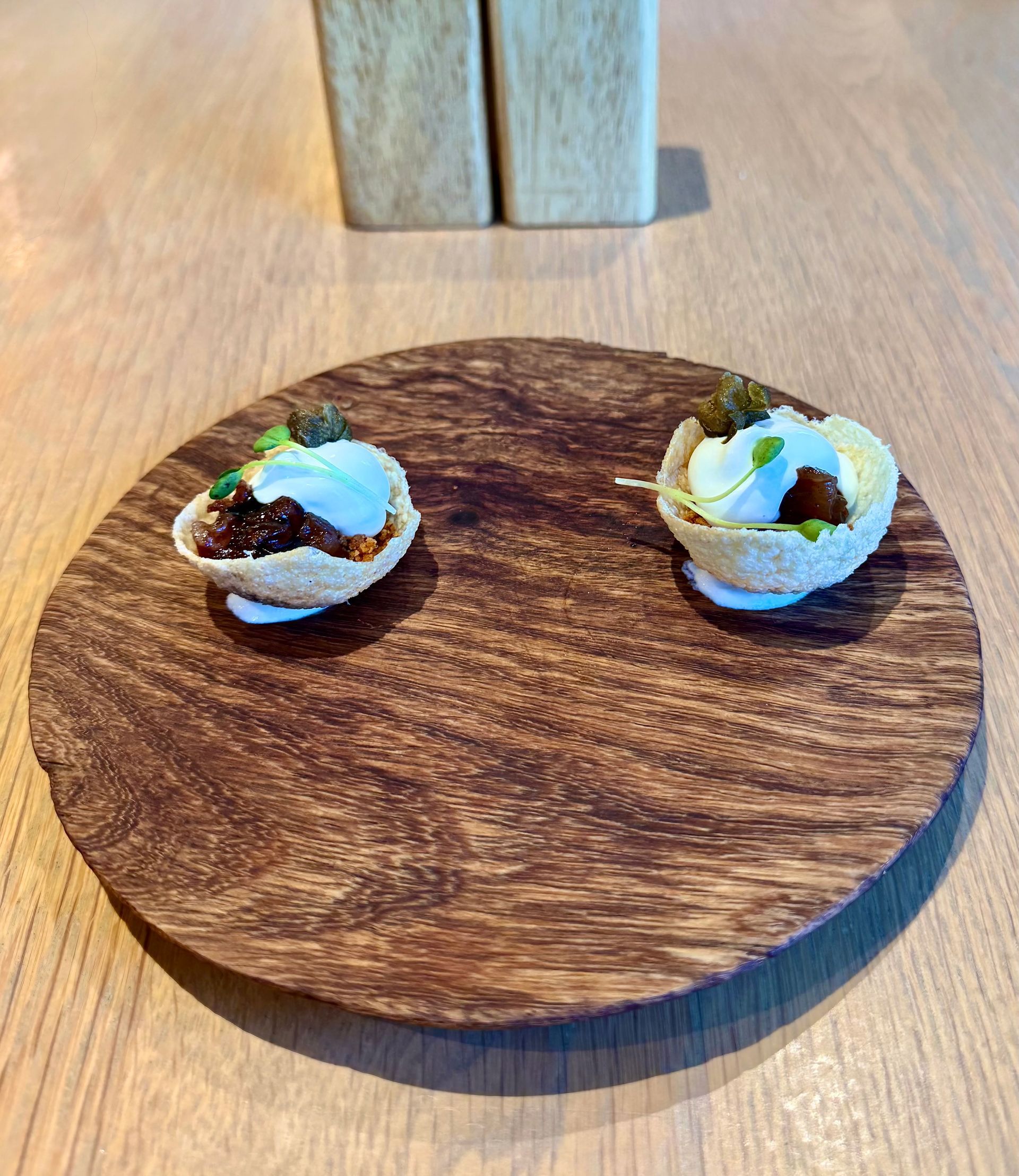 Two small food dishes on a wooden plate, each filled with various toppings.