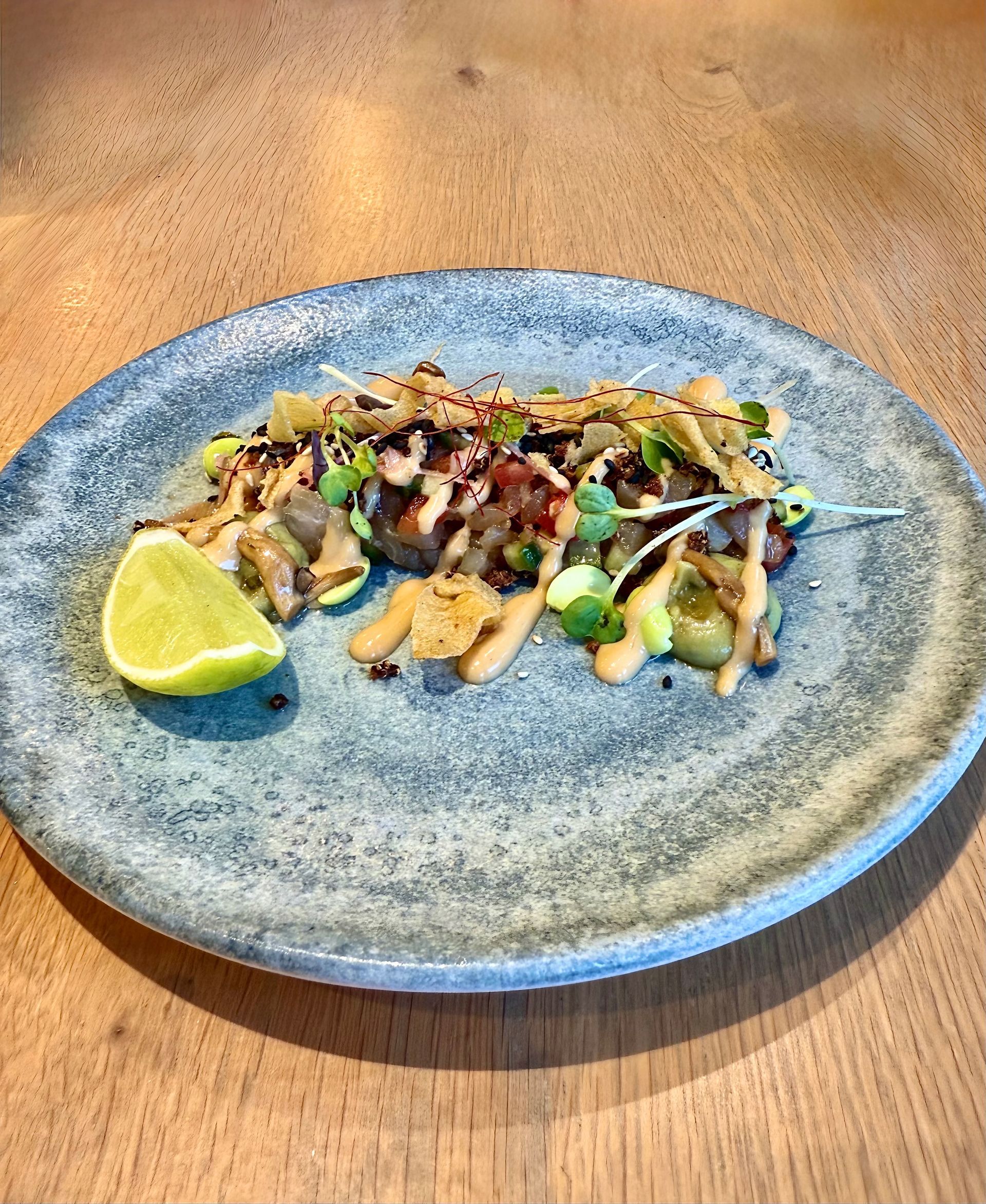 A colorful plate of food, garnished with microgreens, a lime wedge, and drizzled with sauce, on a wood table.