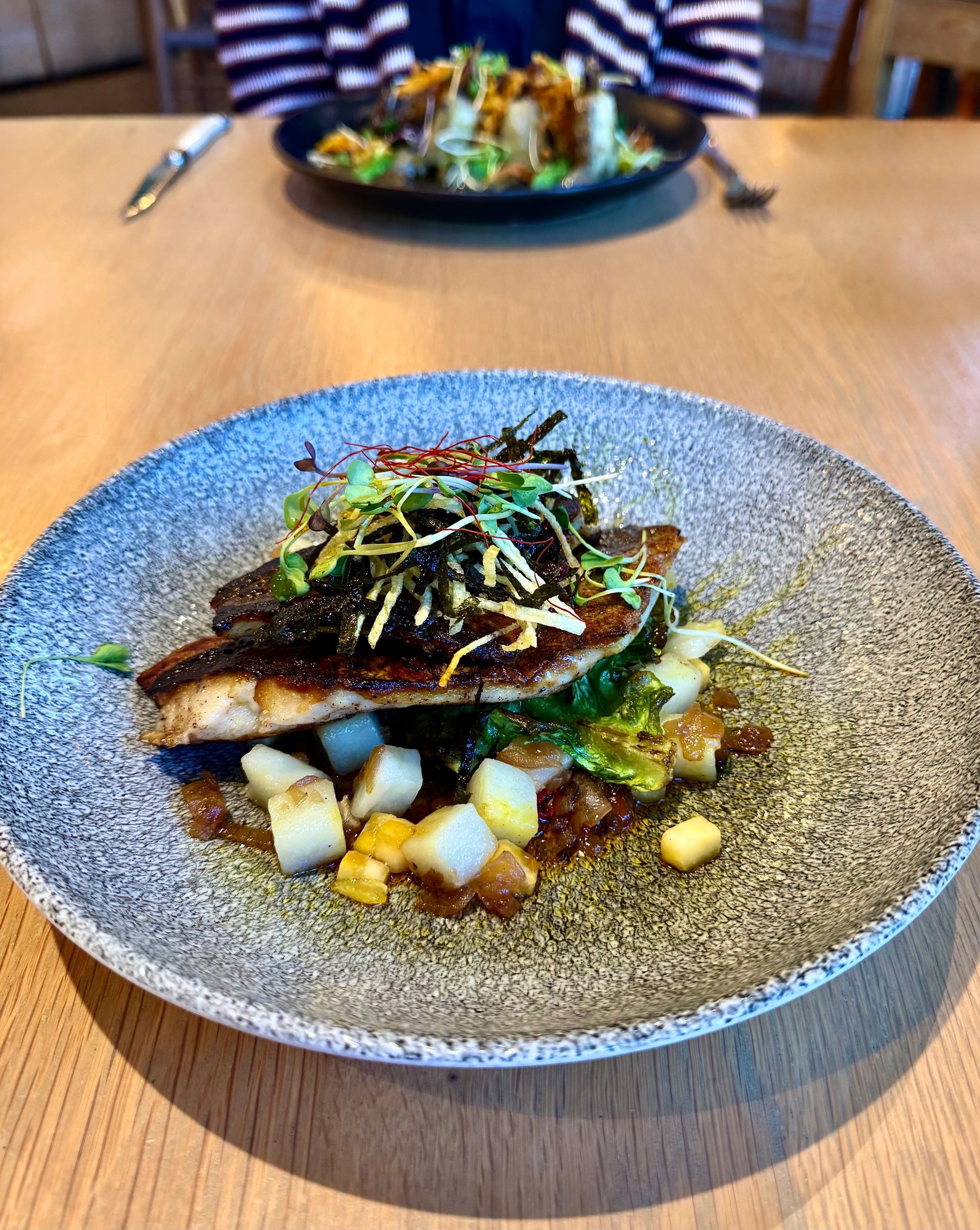 Seared fish on a textured gray plate, garnished with greens, sitting on a table. Another dish in the background.