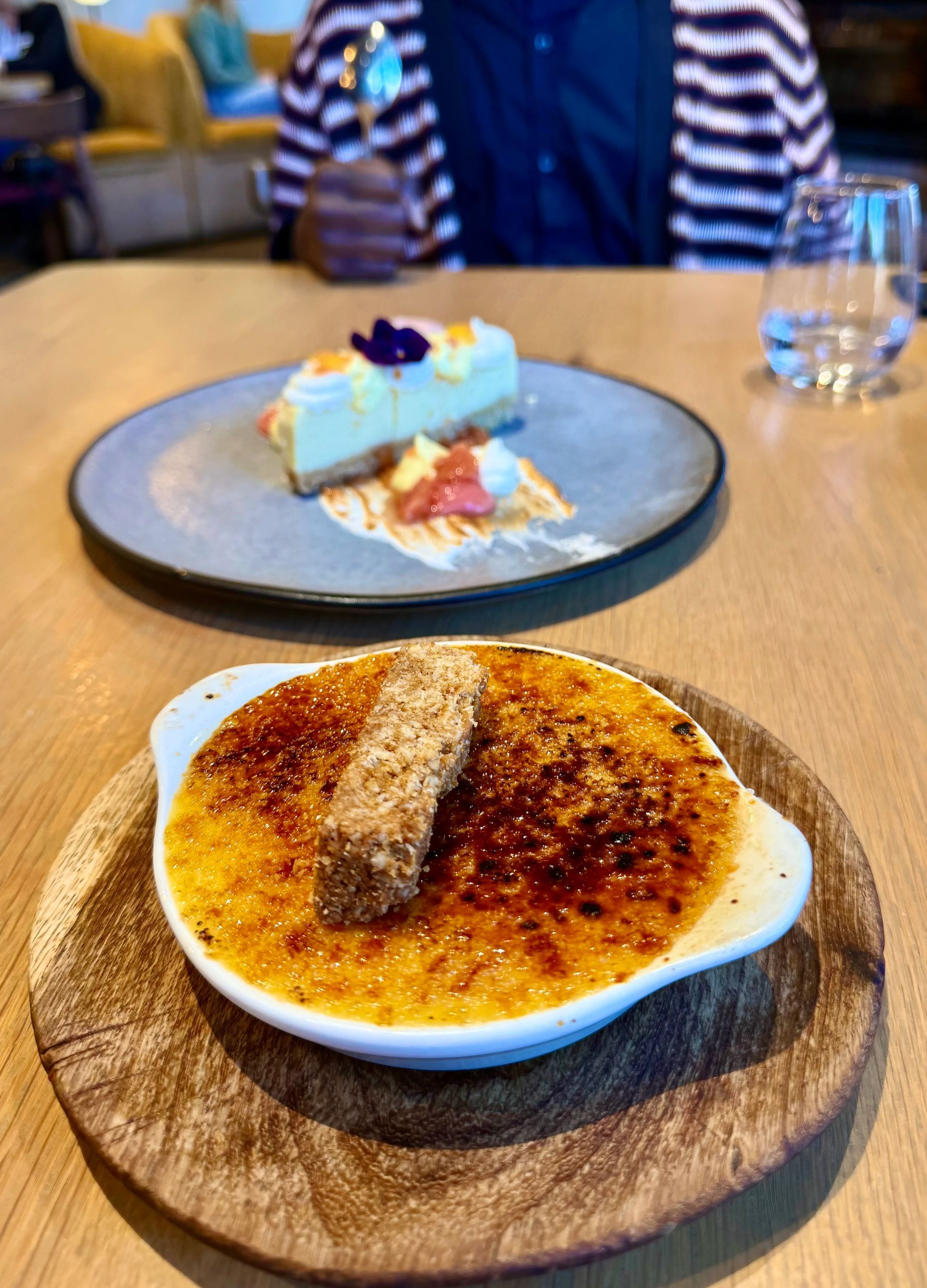 Two desserts on a wooden table: Crème brûlée and cheesecake with a person in the background.