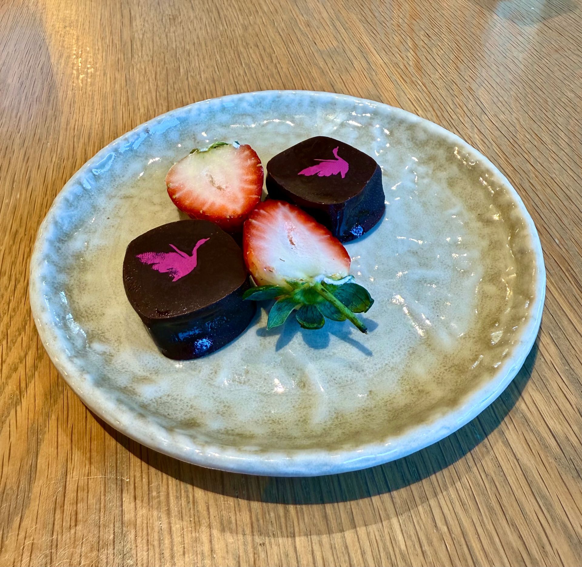 Two dark chocolates topped with a pink crane logo, and two strawberry halves on a small, speckled plate.