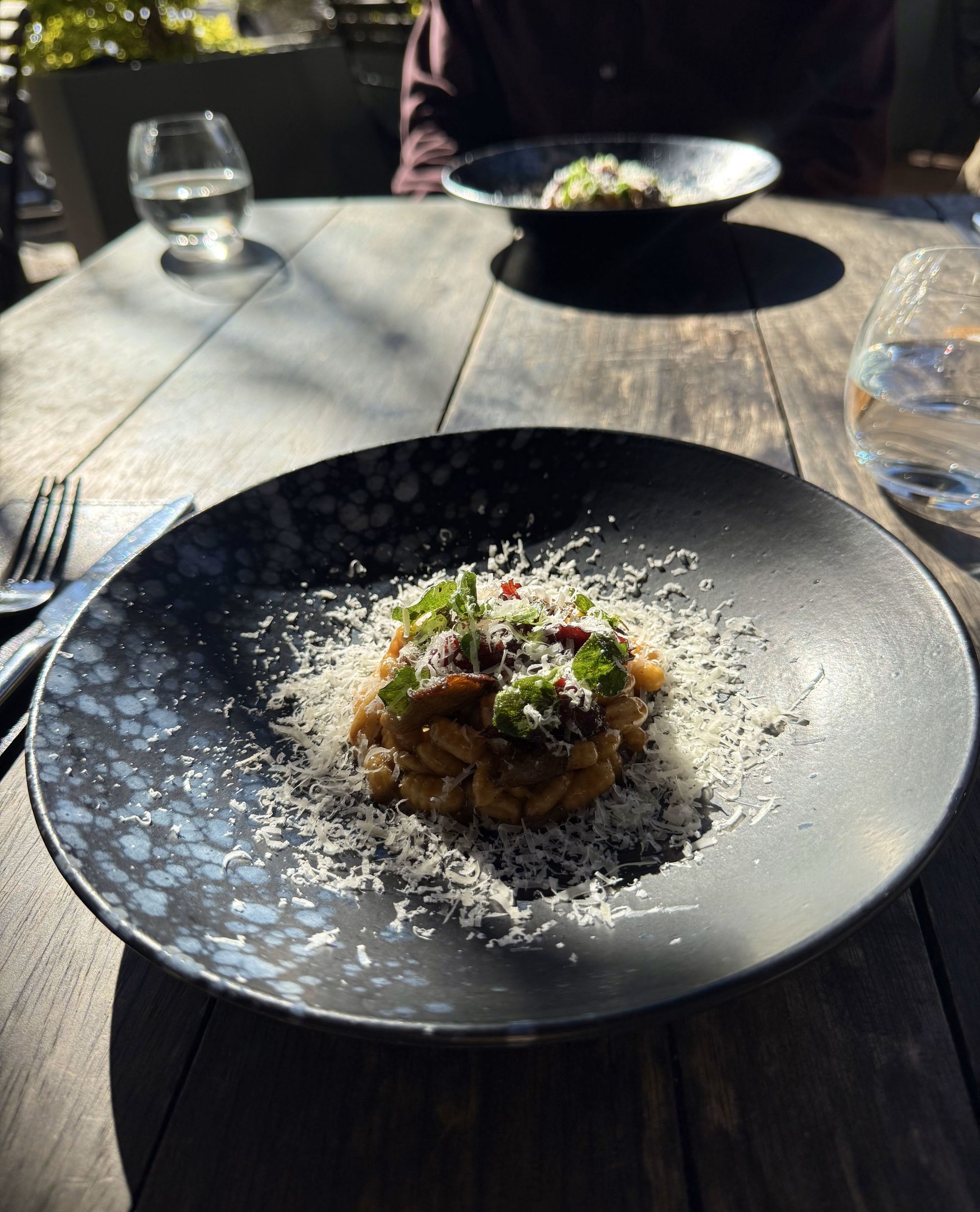 Pasta dish with parmesan on a wooden table, sunlight, and a person blurred in the background.
