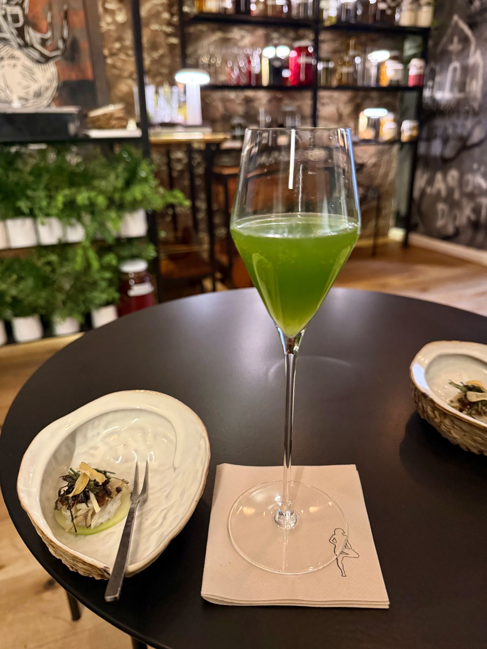 A tall, slender glass of green drink on a black table with a small appetizer and napkin.
