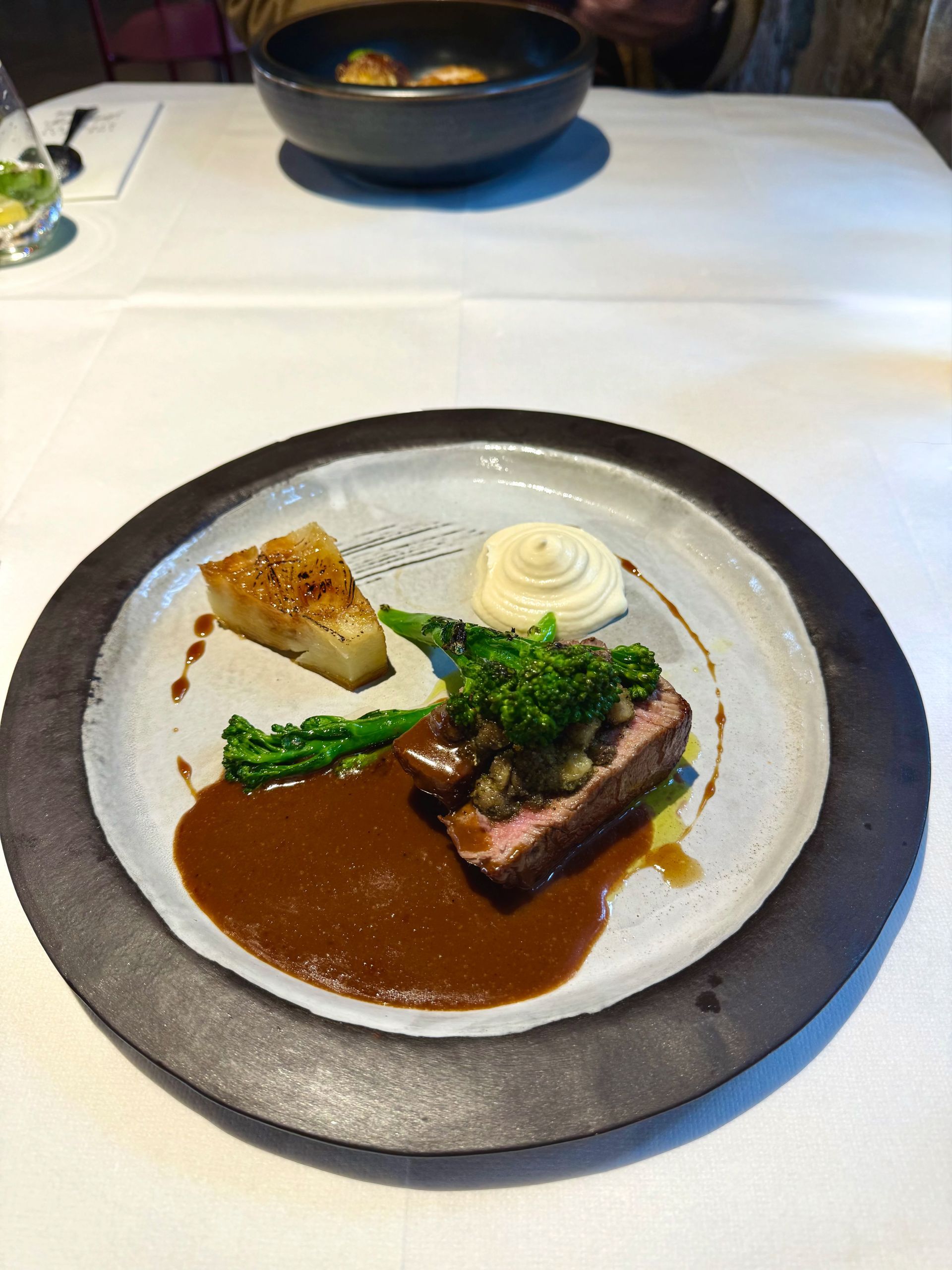 Steak with sauce, potato, and greens on a plate. Dining table set with bowl in the background.