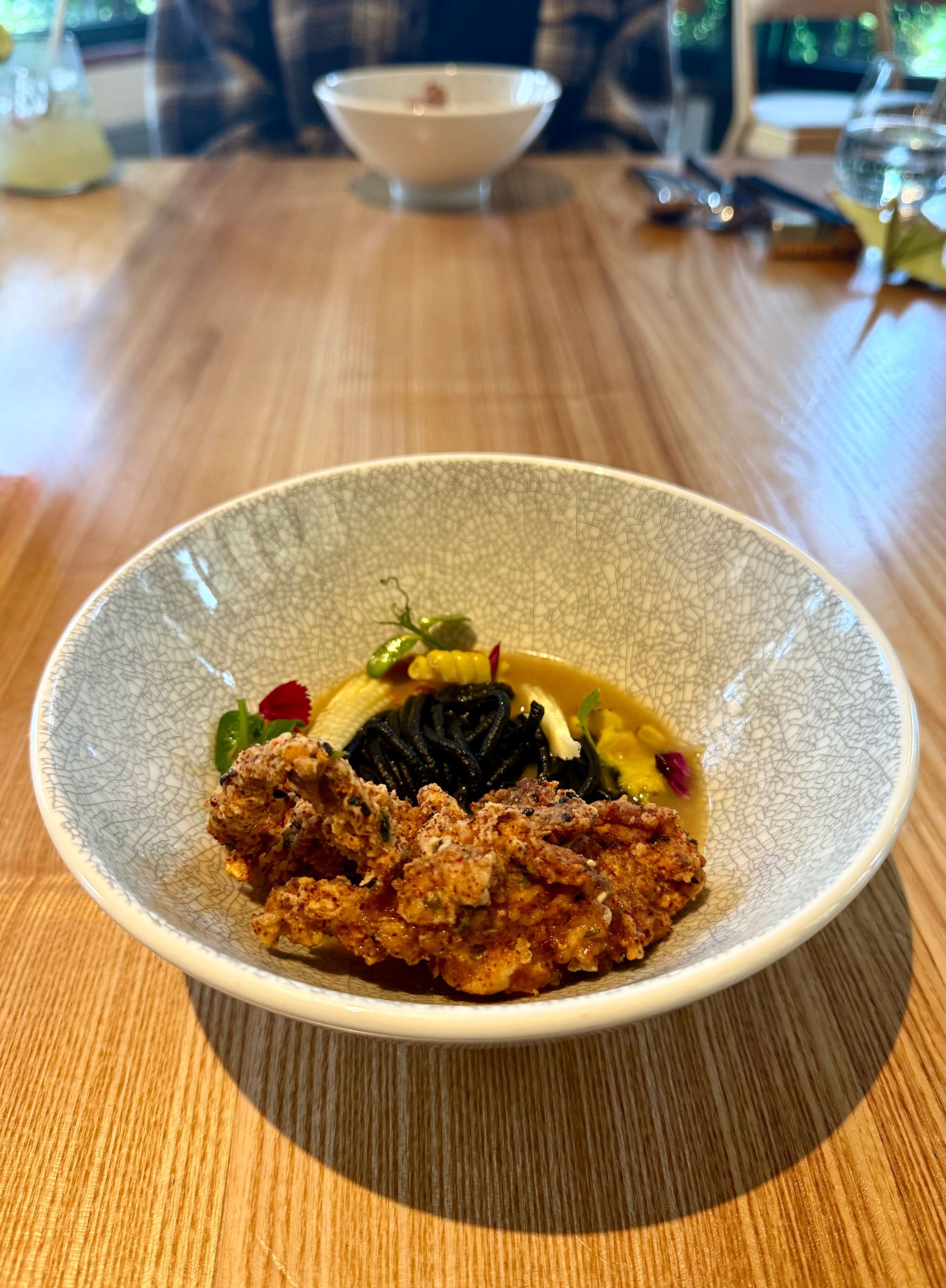A gourmet dish with fried food, sauce, and garnishes in a speckled bowl on a wooden table.