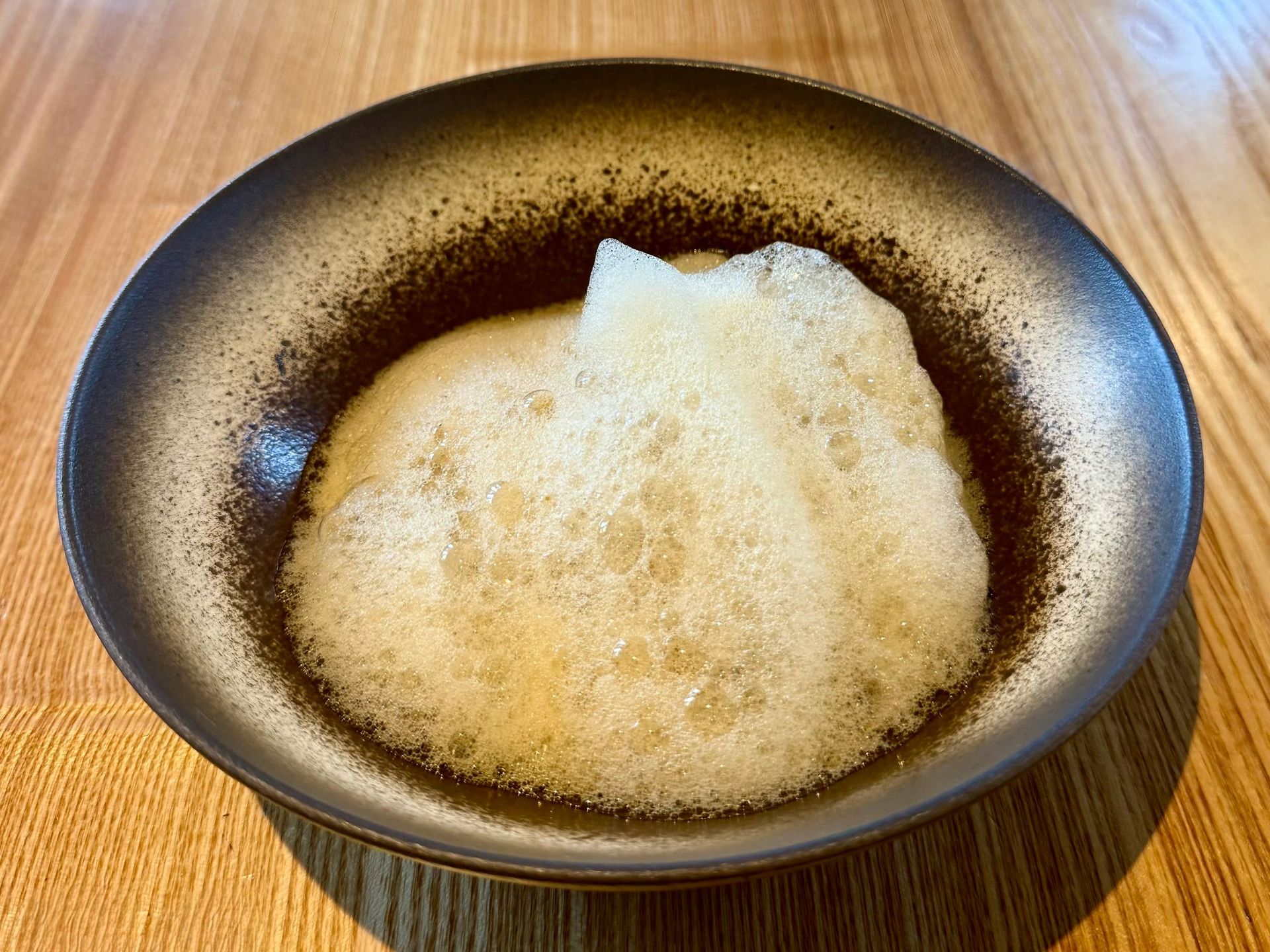A bowl of light-colored foamy soup on a wooden table. The bowl has a dark rim.