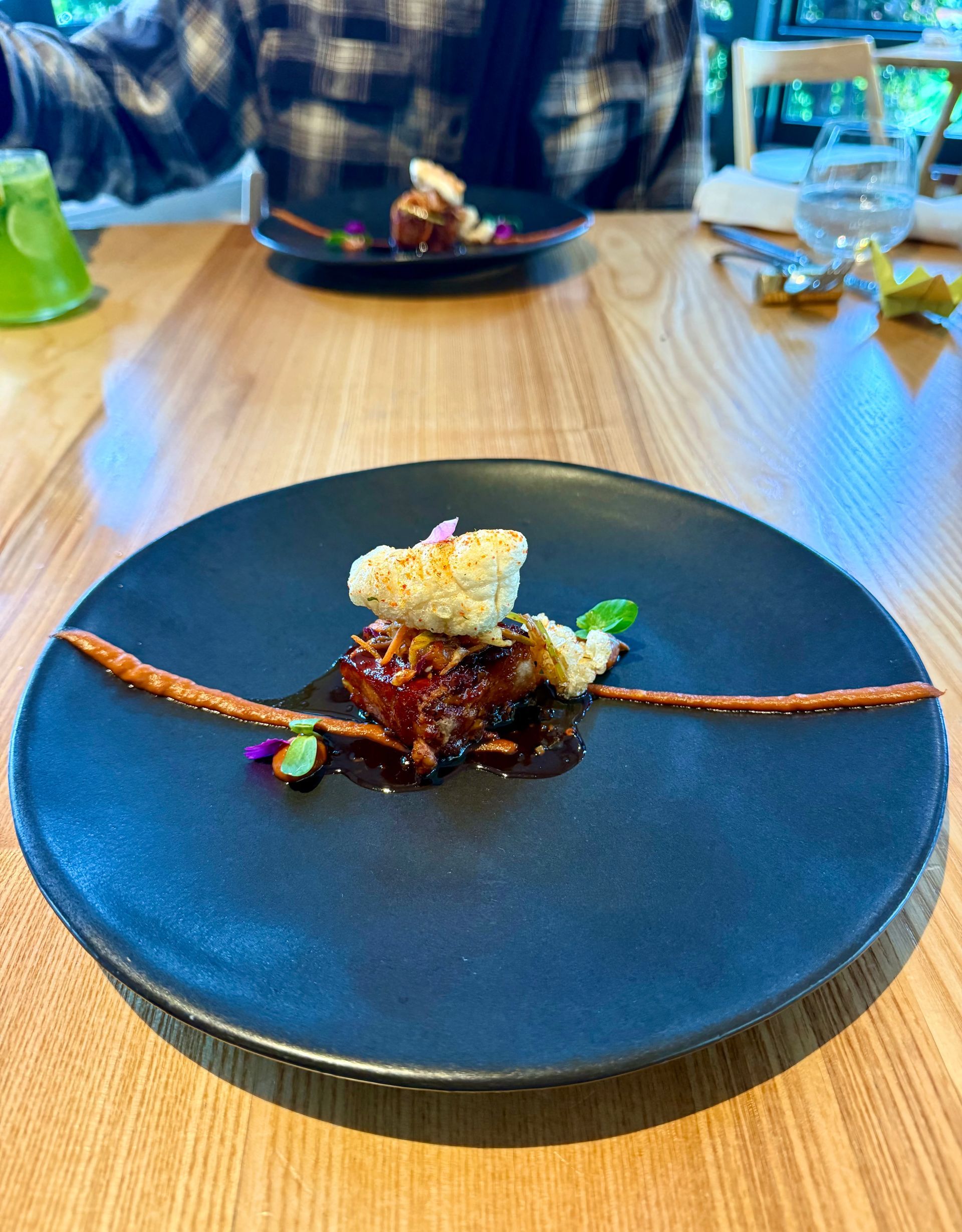 A small dish with food on a black plate, a person in the background, on a wooden table.