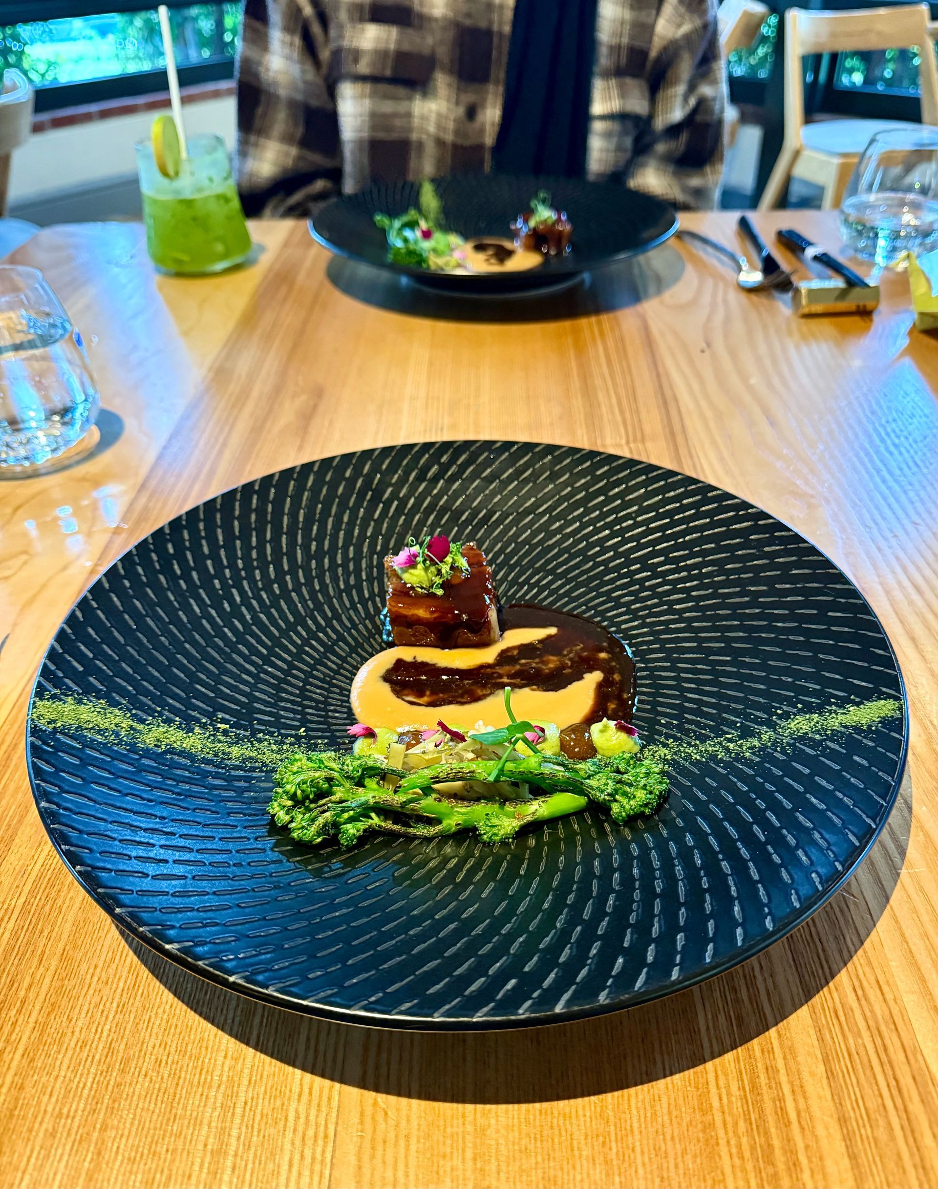 Black plate with gourmet food: seared meat, green sauce, and garnish. A person sits at a table.