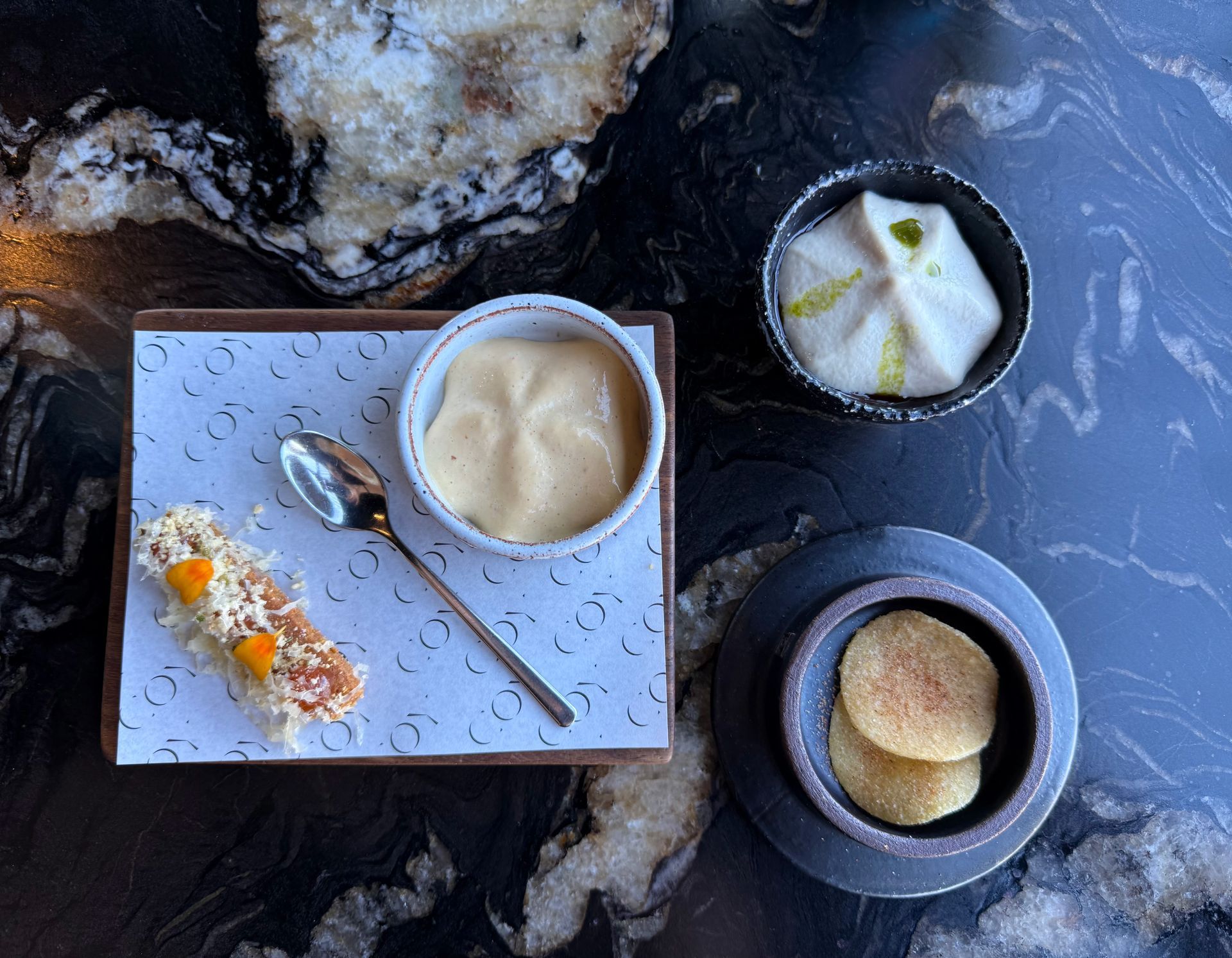 Overhead view of small dishes: a dumpling, a cracker with sauce, a bowl of sauce, and pancakes, on a dark, patterned surface.