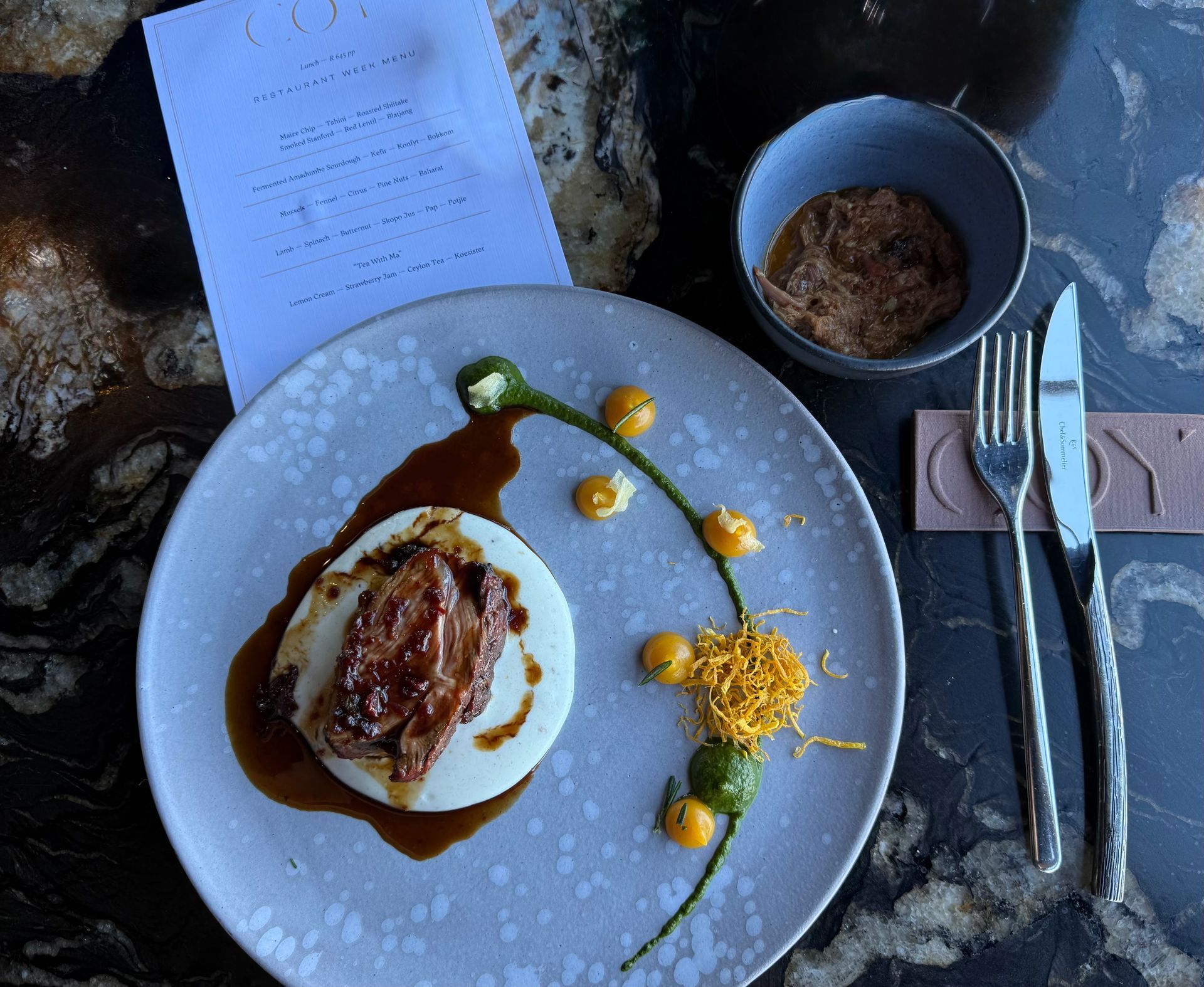 A plate with meat, sauces, and decorative garnishes beside a bowl of sauce and silverware on a table.