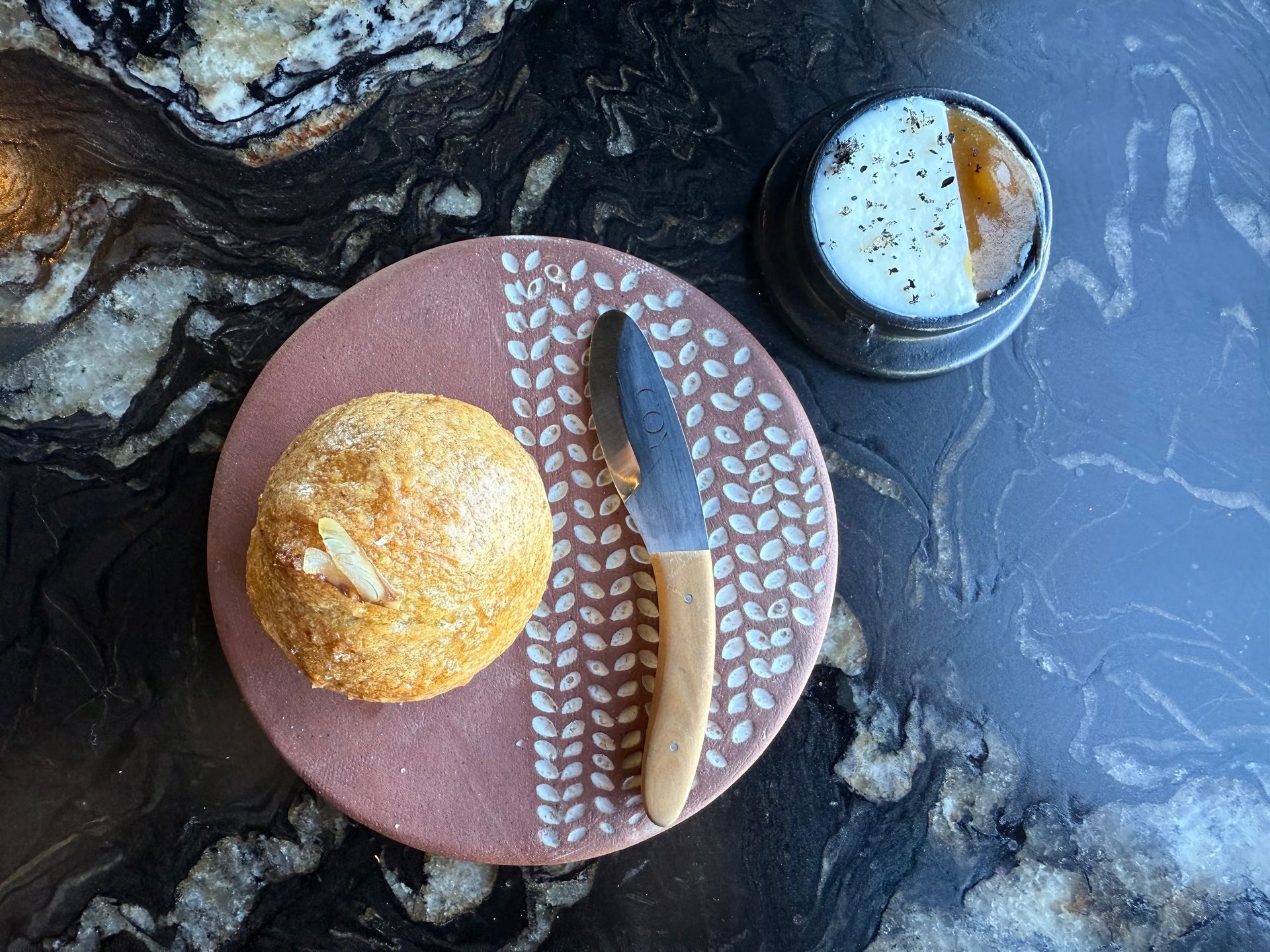 Bread roll with butter and knife on a patterned pink plate next to a butter dish on a dark countertop.