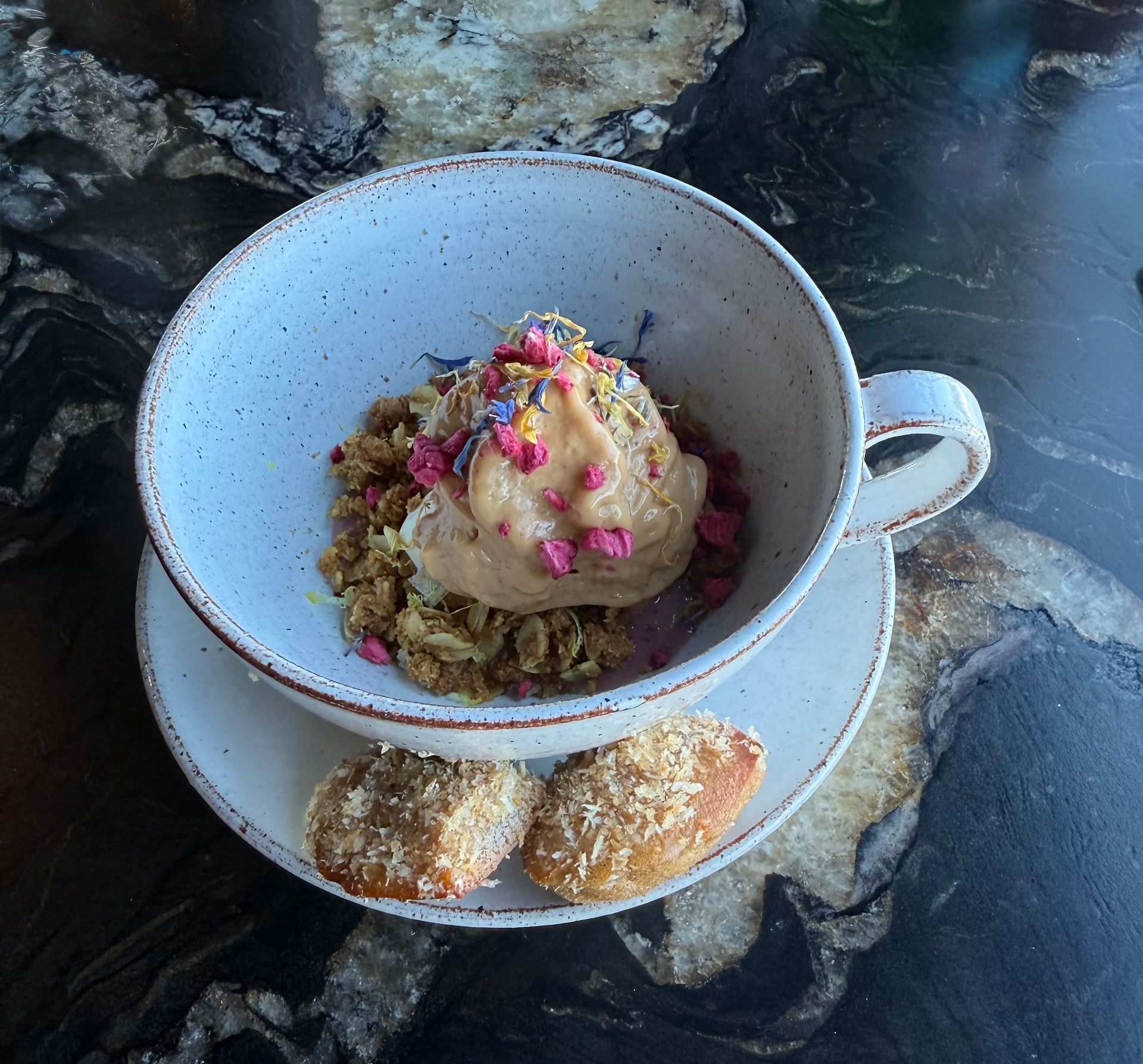 Dessert in a speckled cup with granola, ice cream, and flower petals, served with two pastries on a saucer.