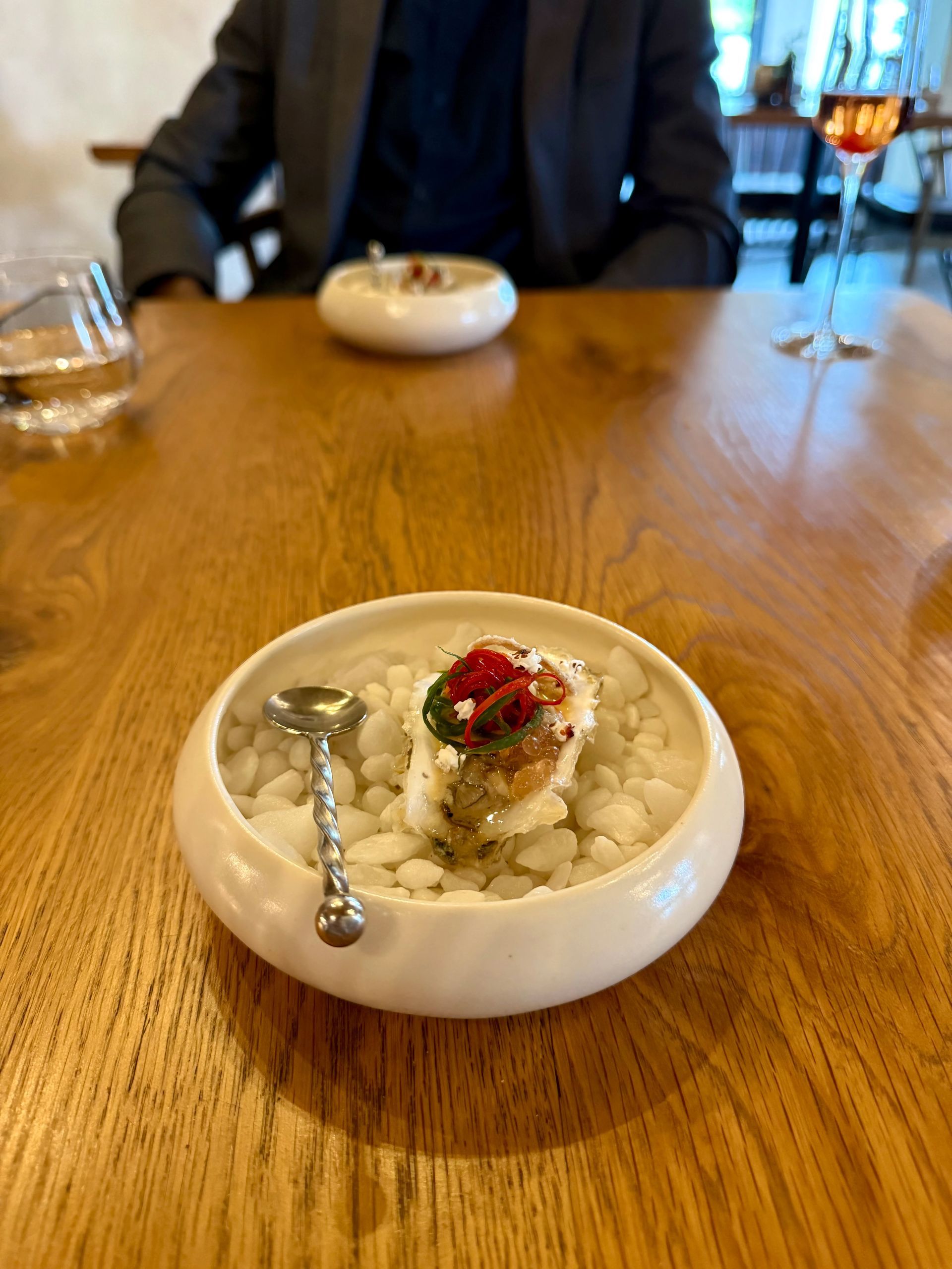 Small dish with food and spoon on a wooden table; person seated in background.