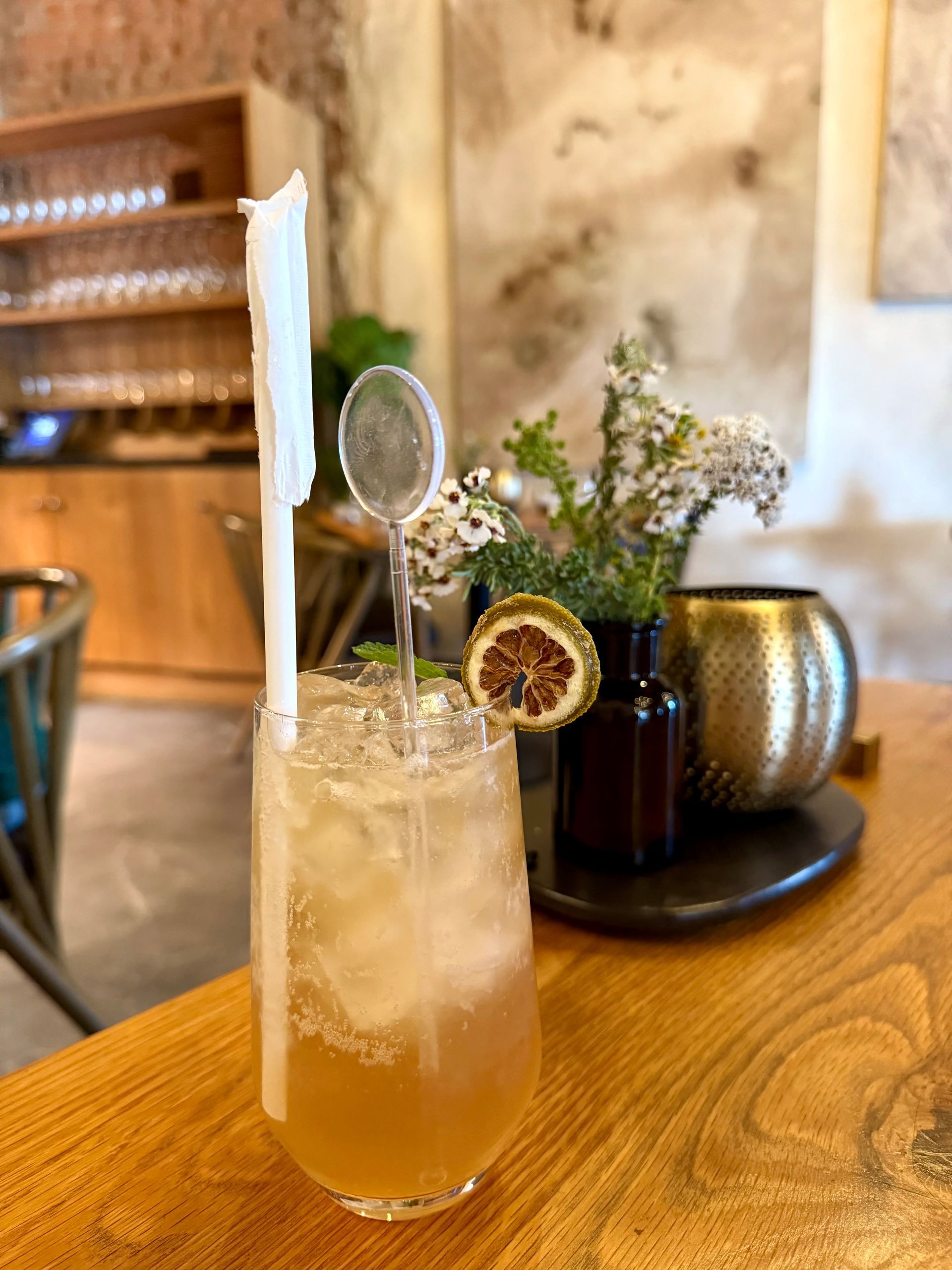 Tall cocktail with ice, garnish, and spoon straw, on a wood table. Background features a vase of flowers.