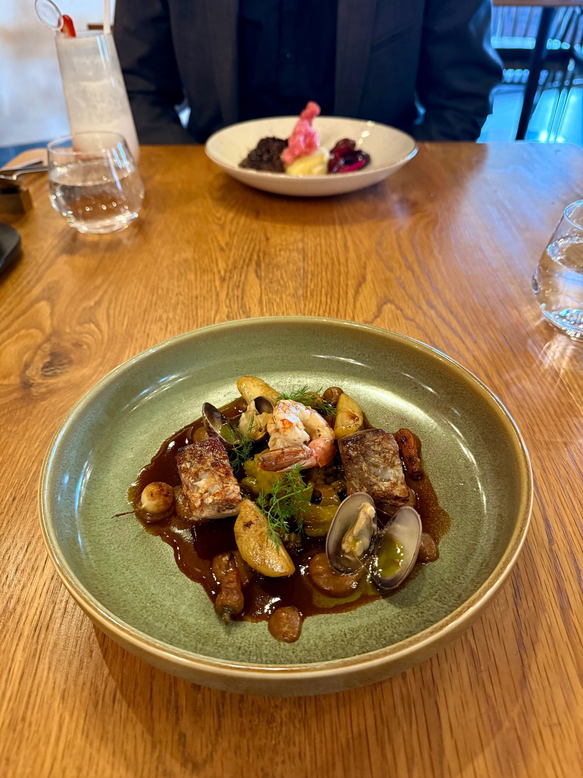 Plate of food with meat, vegetables, and sauce on a wooden table, with another dish and person in background.