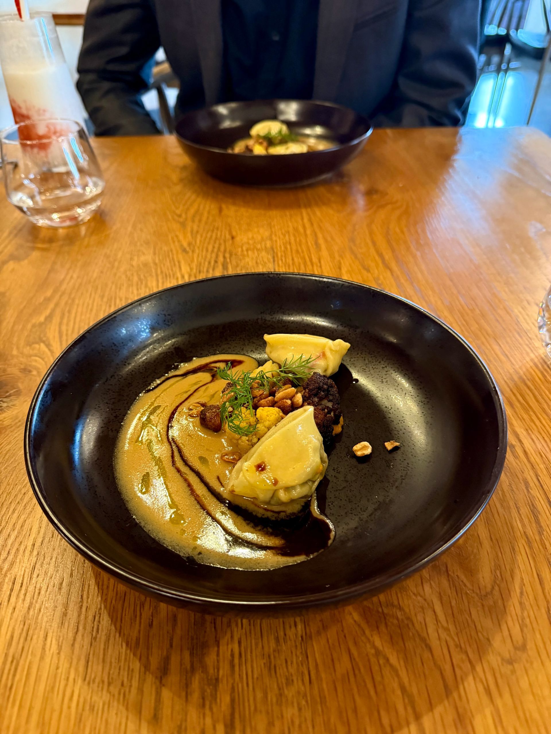 A dark plate of food with sauce, next to another bowl on a wooden table, person in the background.