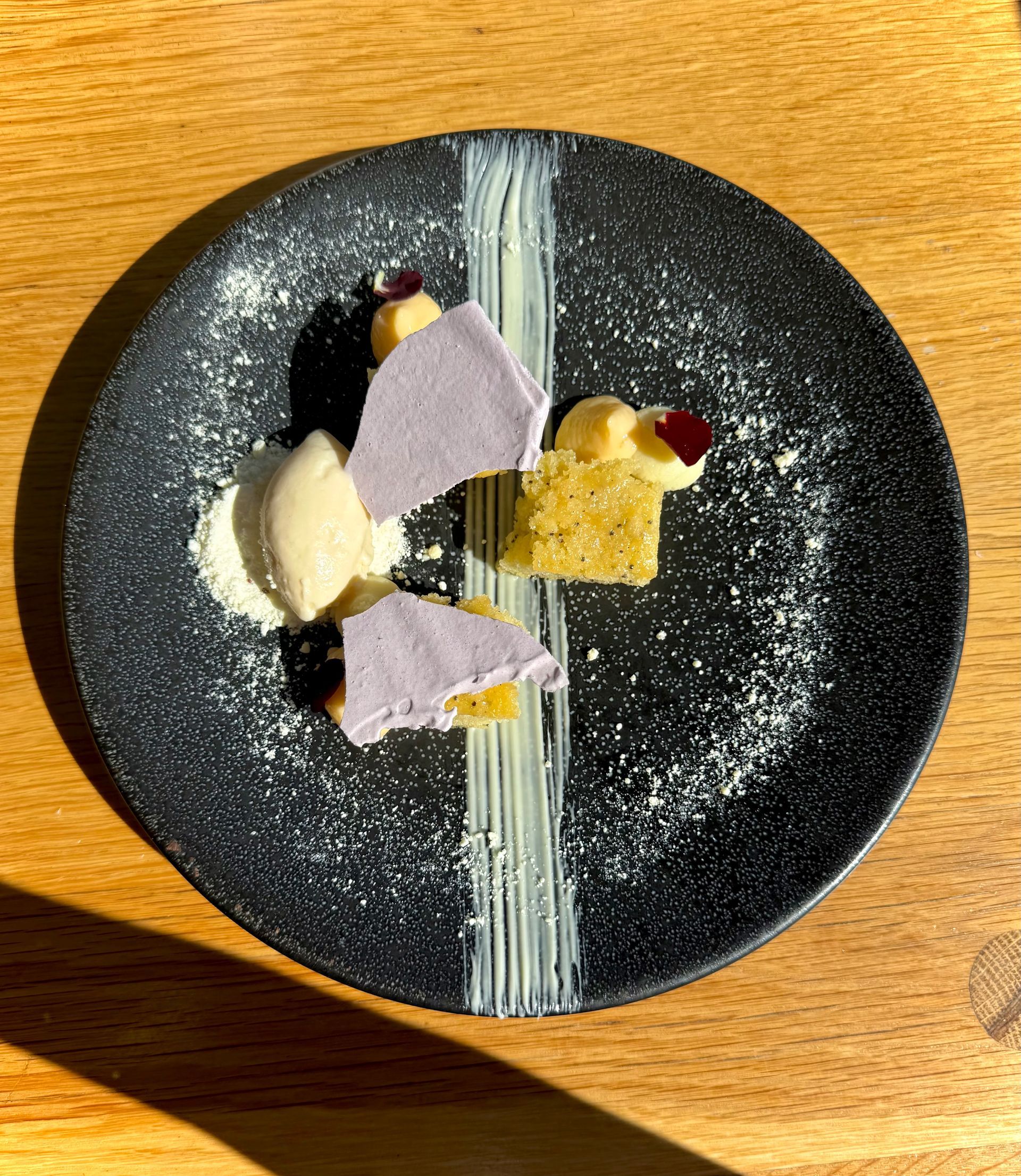 Dessert on black plate: lavender, yellow, and cream elements, powdered sugar, white line.