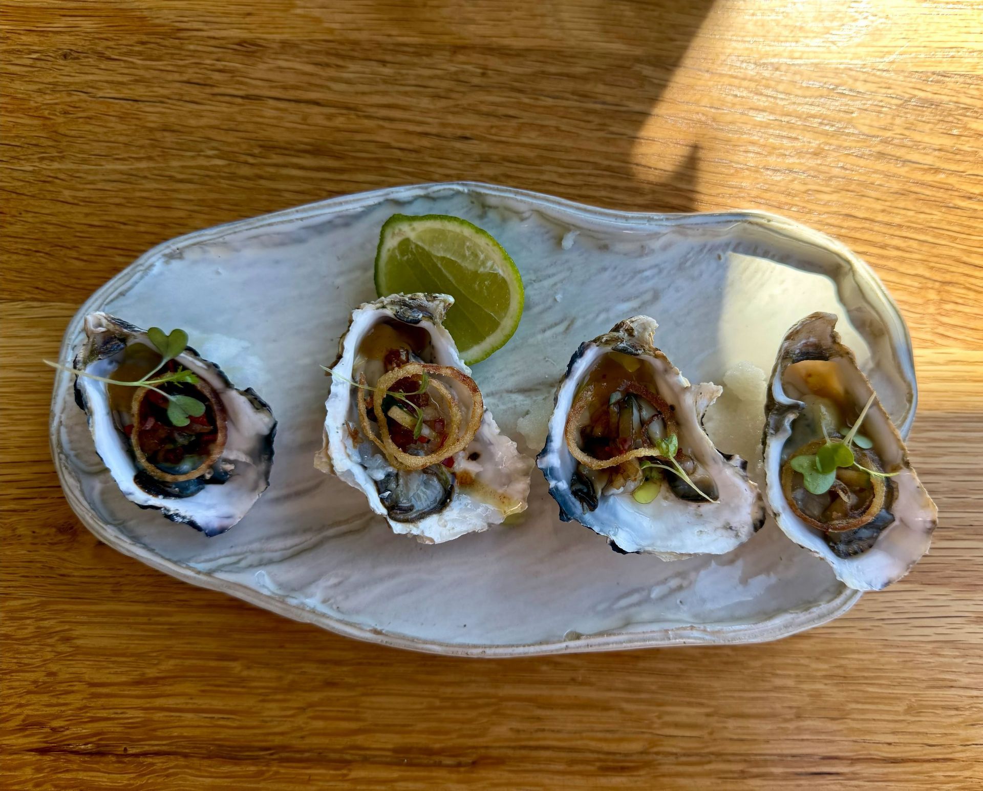 Four oysters on a decorative plate, each with a filling and garnish, served with a lime wedge on a wooden table.