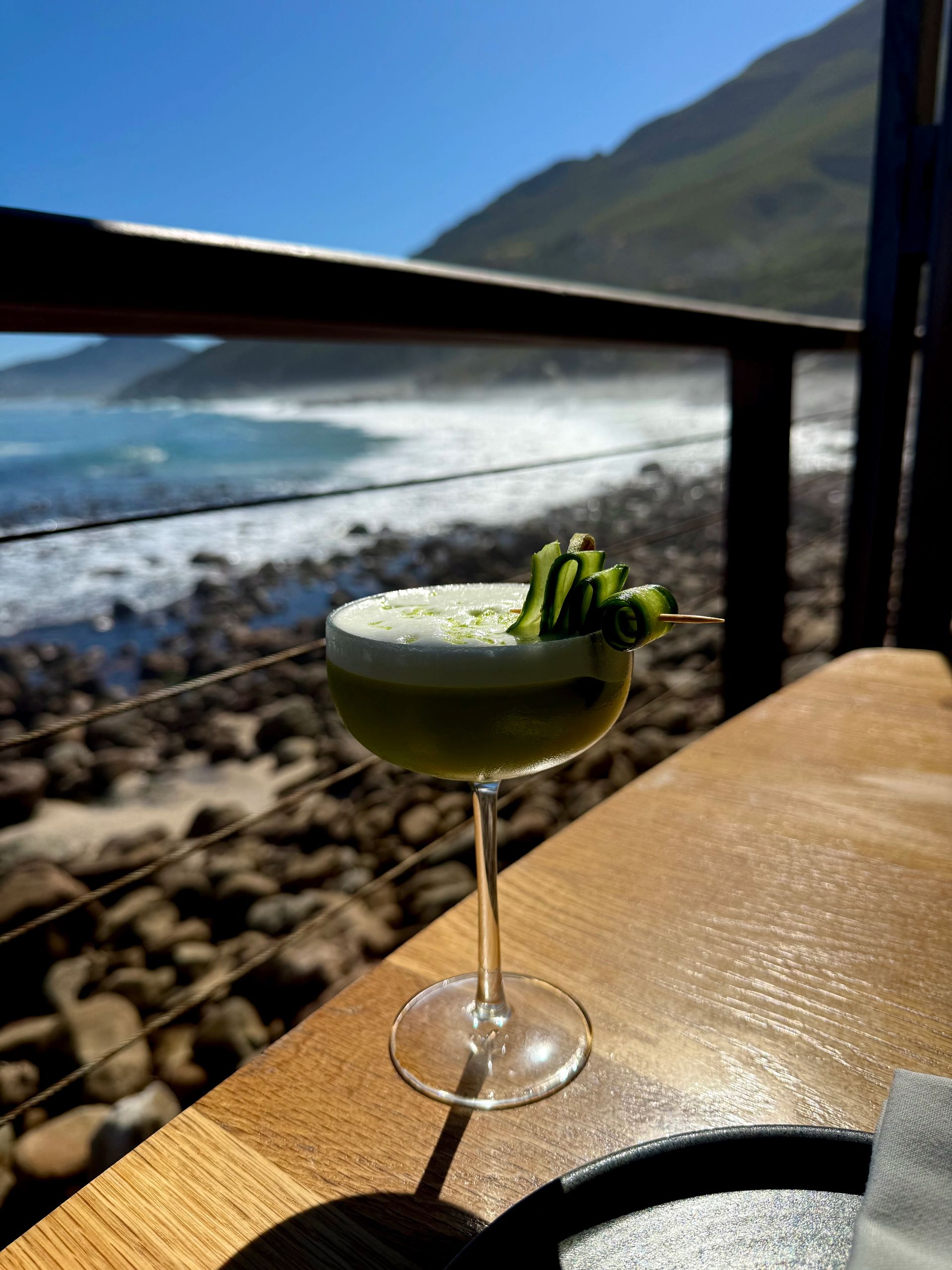 Cocktail on a wooden table overlooking a rocky beach, ocean, and mountains. Green drink with garnish, sunny day.
