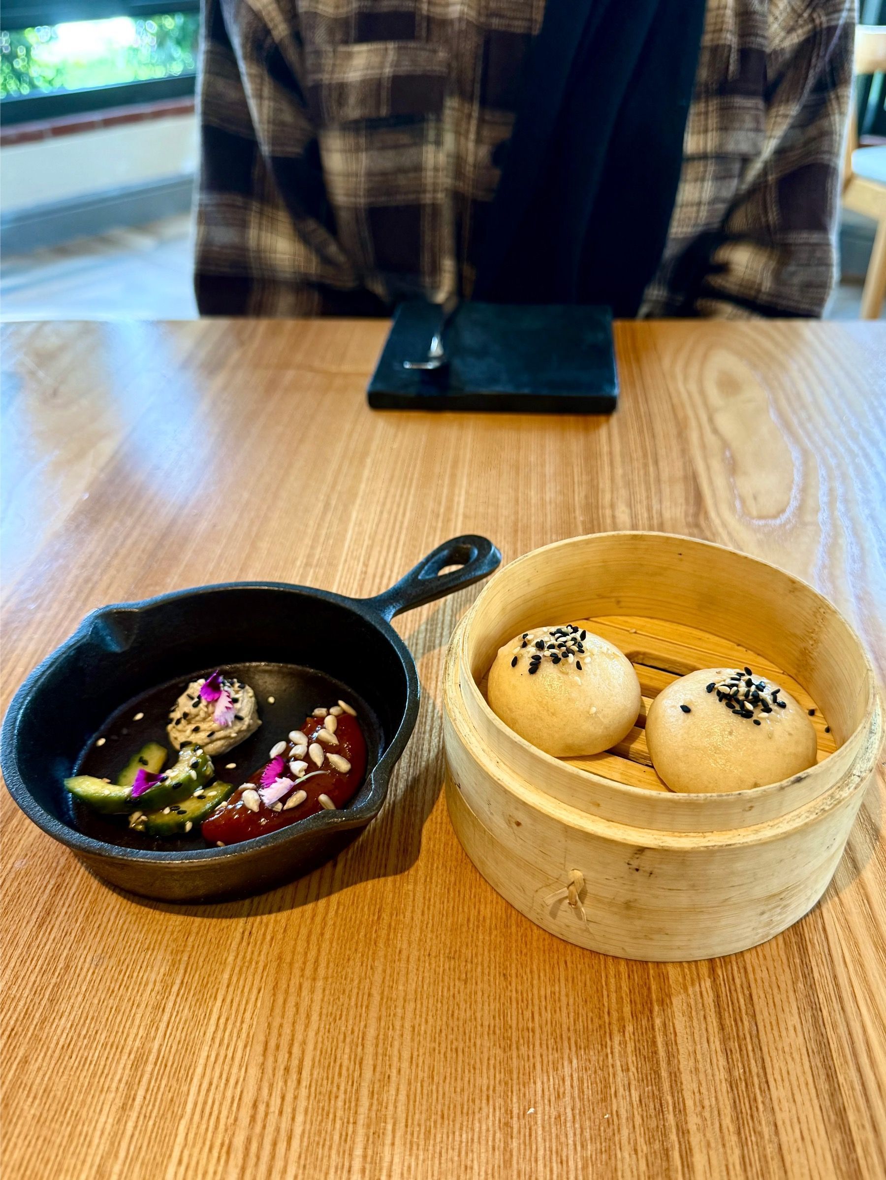 Small cast iron dish with dipping sauce, and a bamboo steamer with two buns on a wooden table.