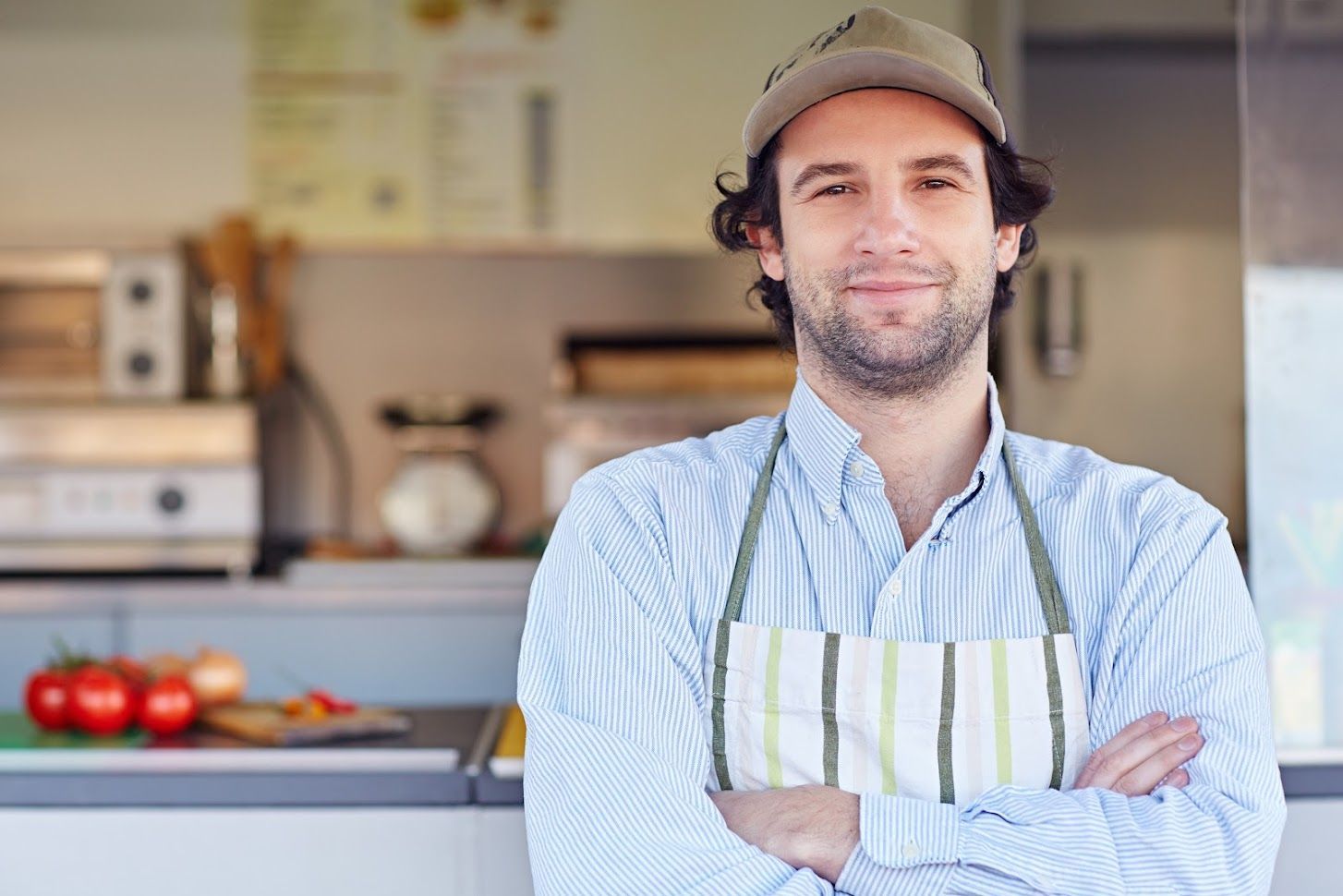A man wearing an apron and hat is standing in a kitchen with his arms crossed.
