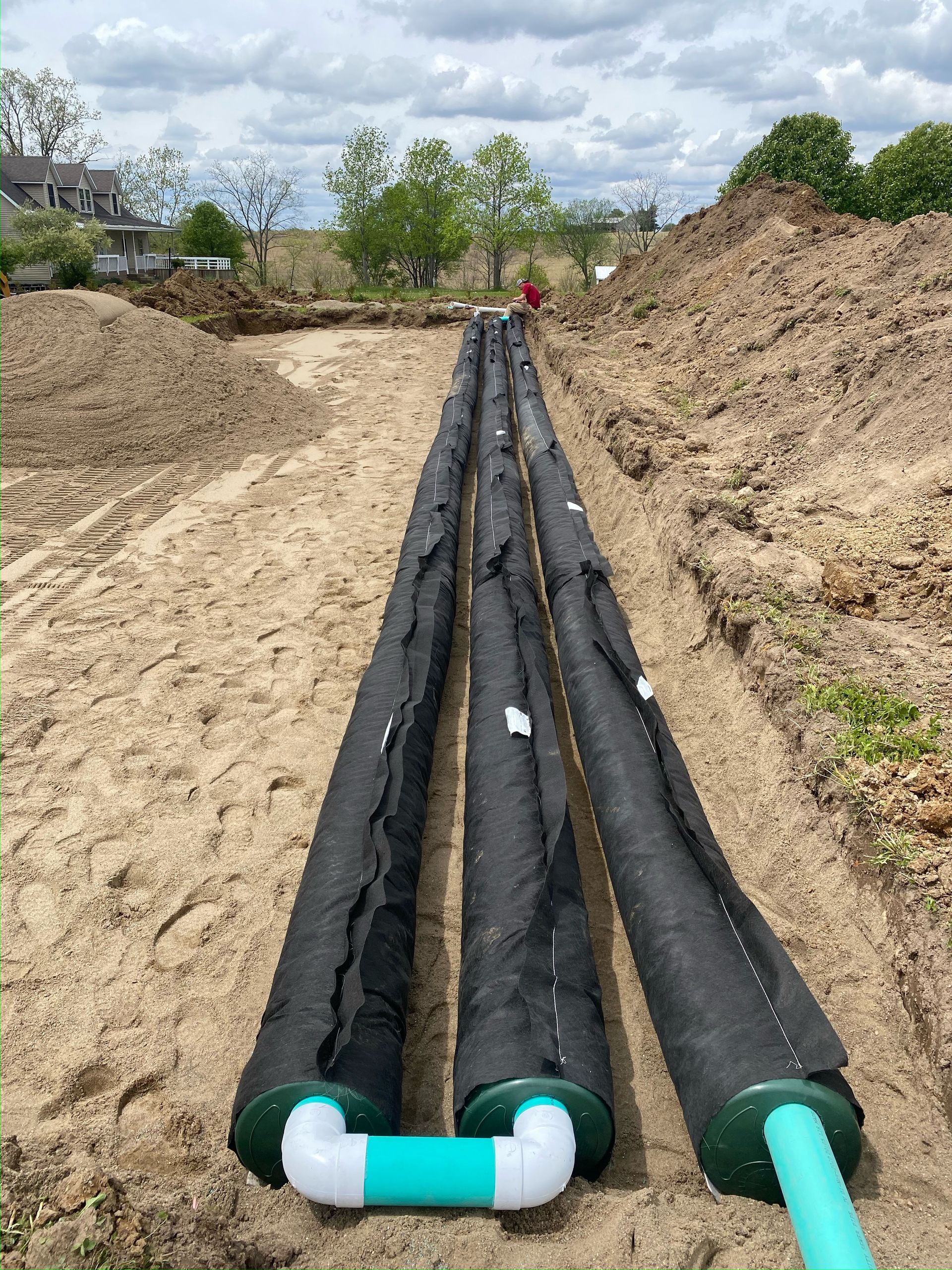 Three black wrapped drain pipes in a trench, connected by green and white plumbing, in a dirt setting.