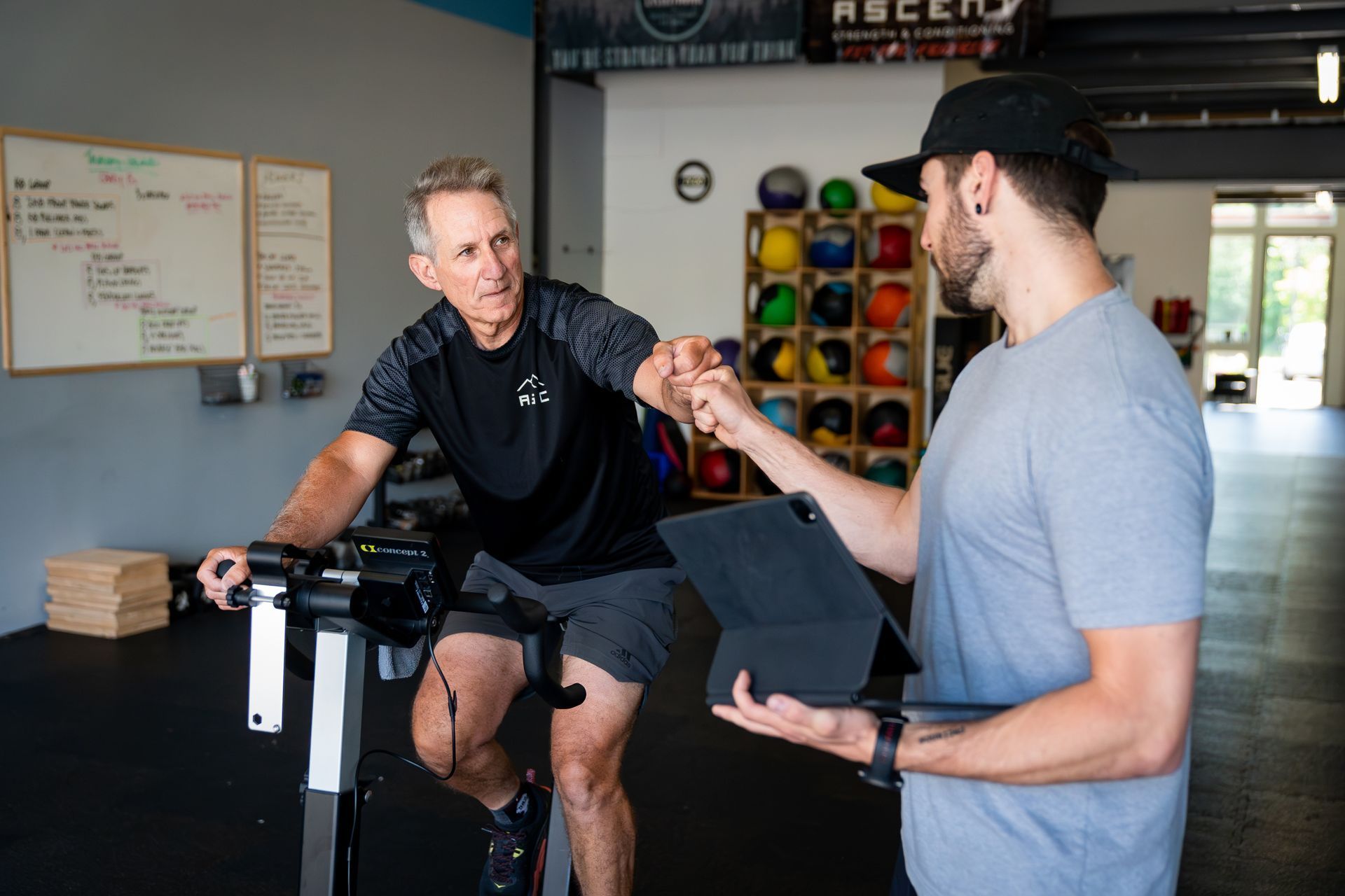 Man on exercise bike bumps fists with trainer holding a tablet in a gym.